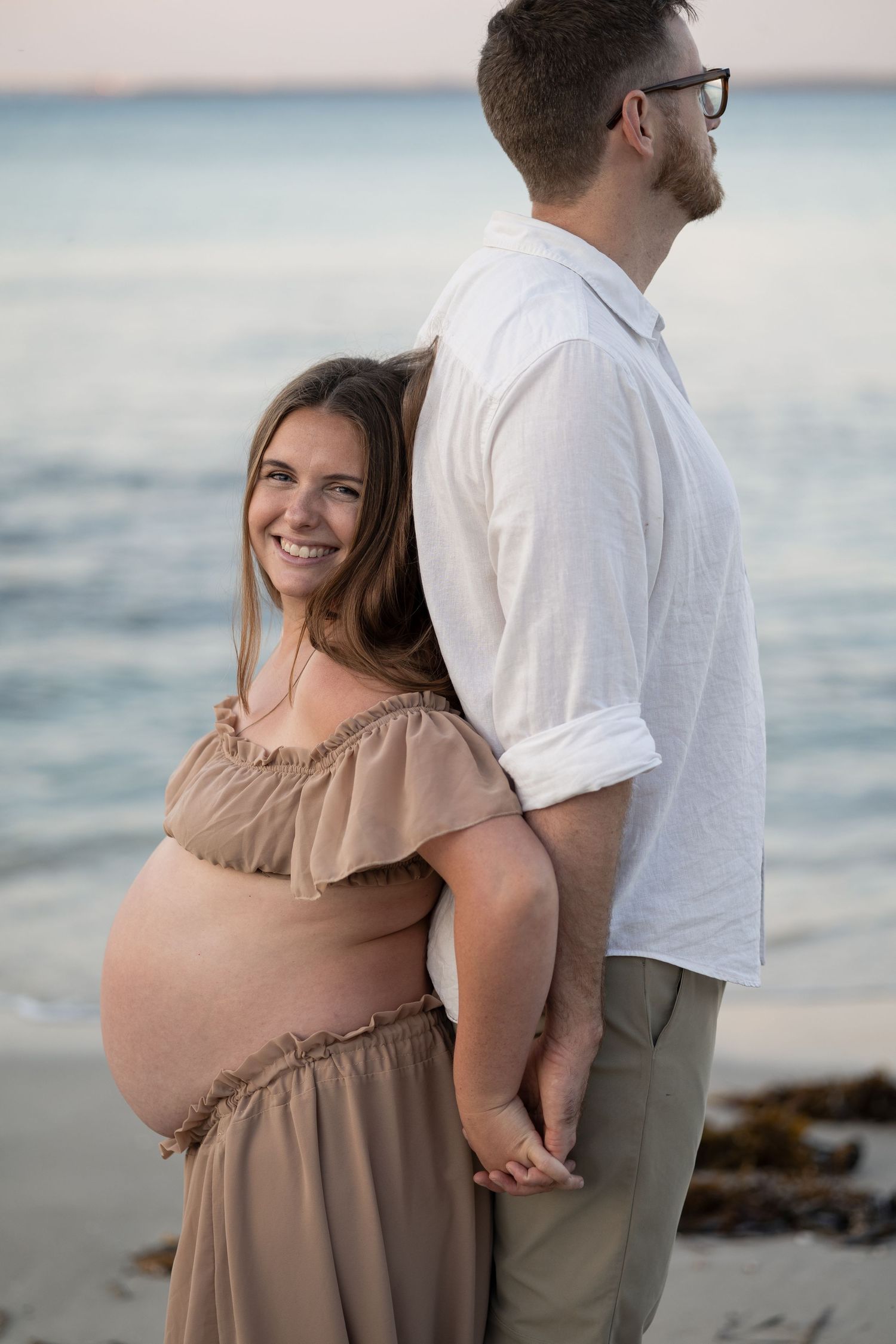 Smiling pregnant woman in beige dress holds hands with man in white shirt on a serene beach at dusk.