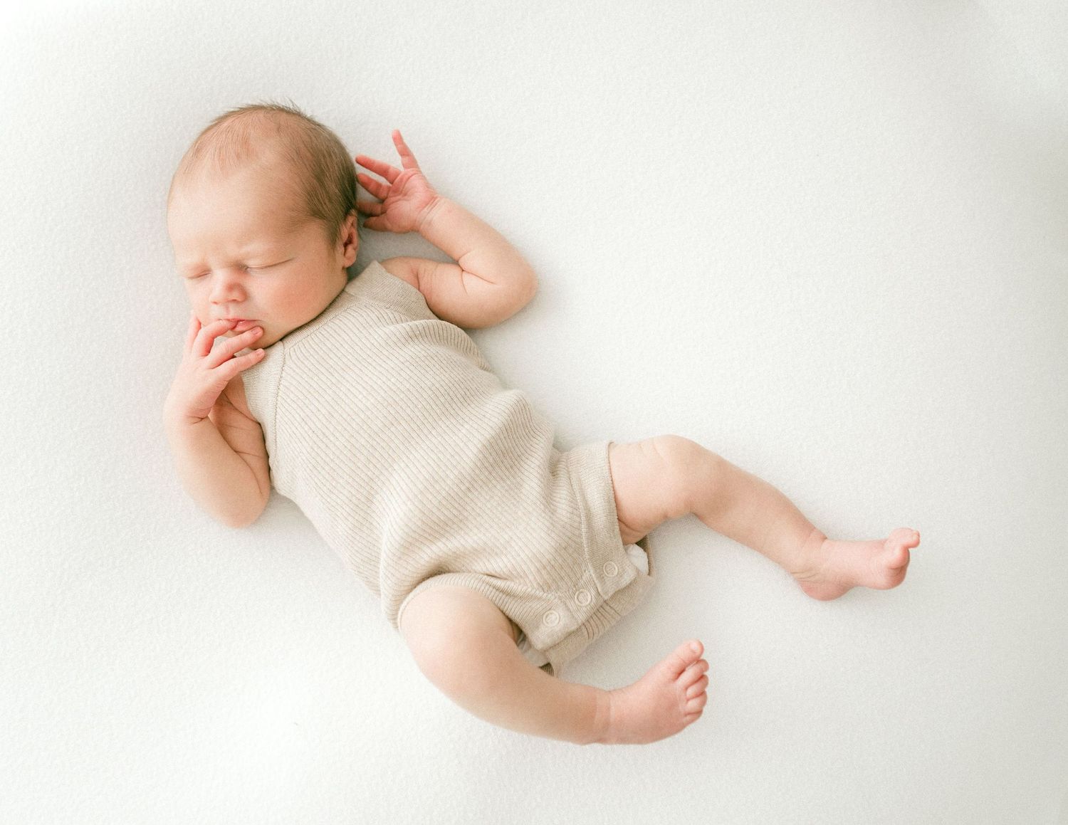 Sleeping newborn baby in beige onesie lying on a white background in a peaceful curled pose.