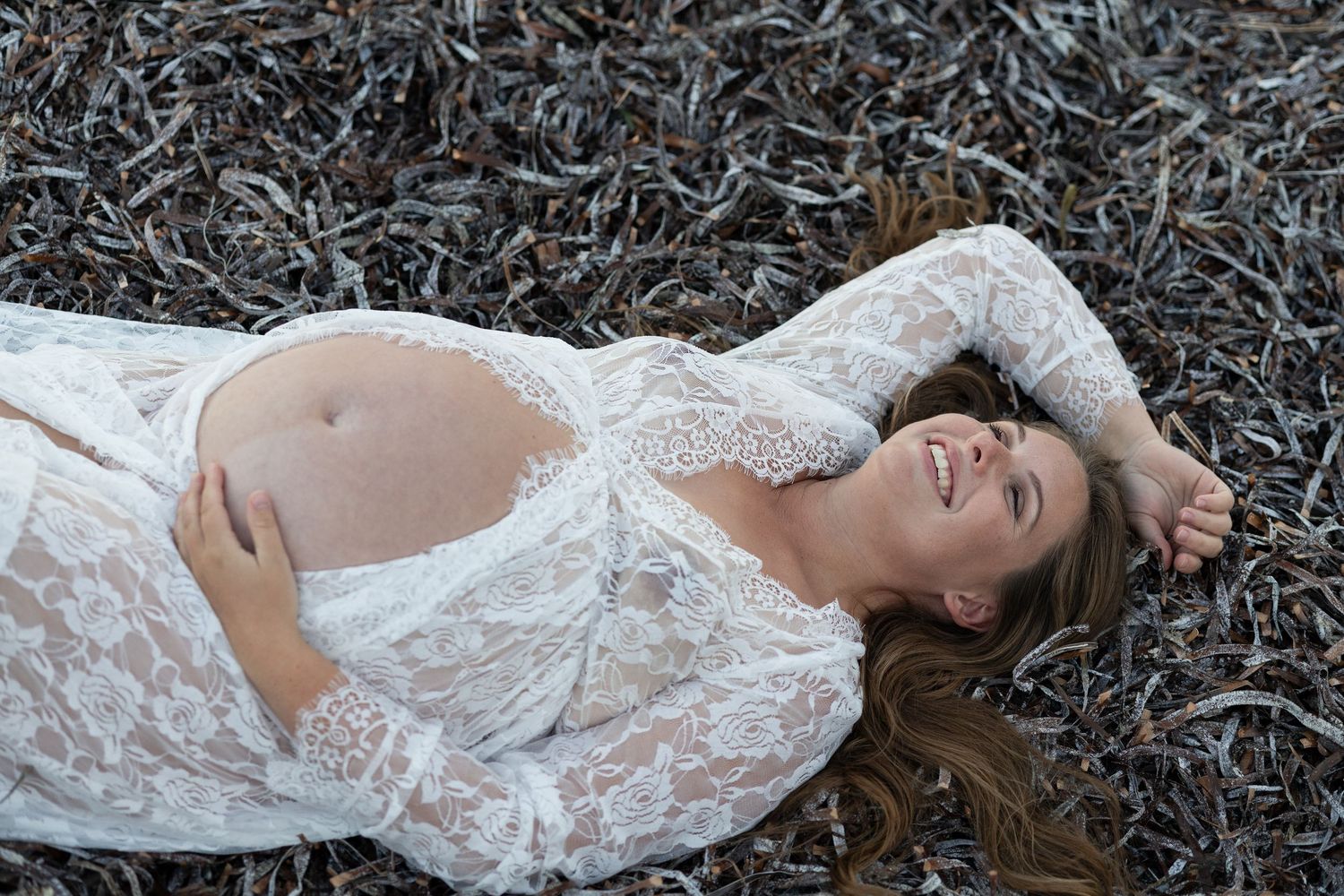 Pregnant woman in white lace dress lying on ground, smiling joyfully during outdoor maternity photo shoot.