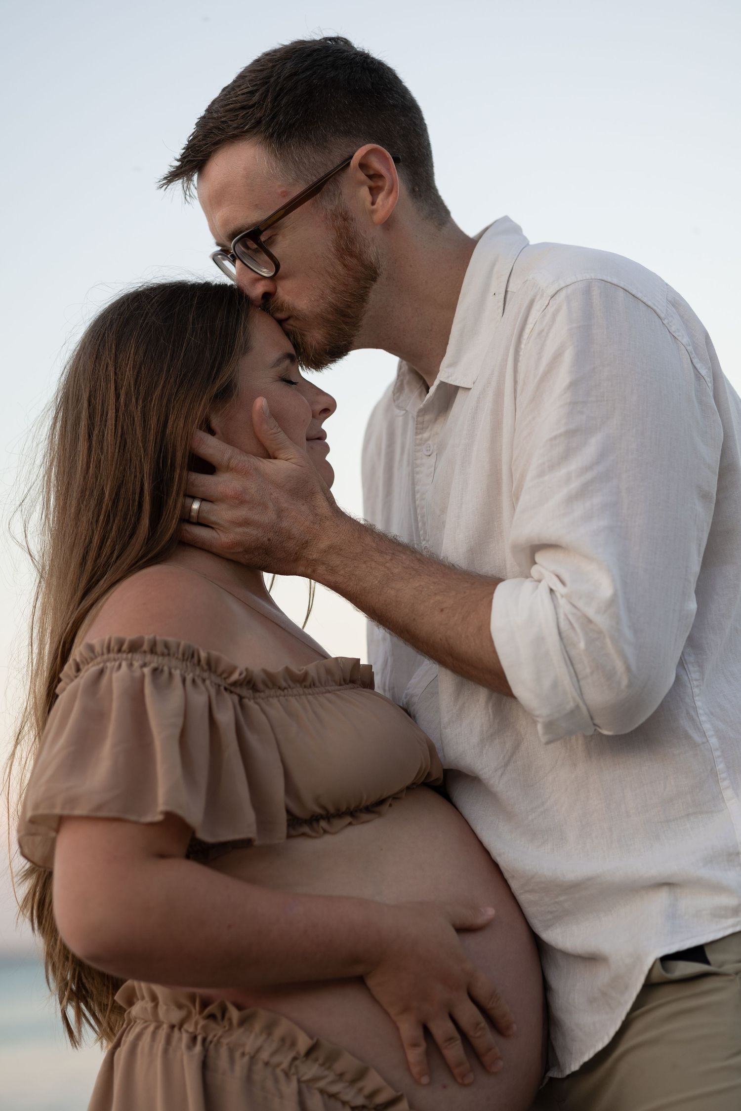 Couple sharing a tender moment on the beach, man kissing pregnant woman's forehead while cradling her belly.