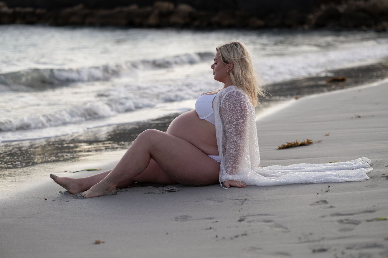 Pregnant woman in white bikini and lace cover-up sitting on sandy beach by ocean waves during golden hour. Woodman Point Perth