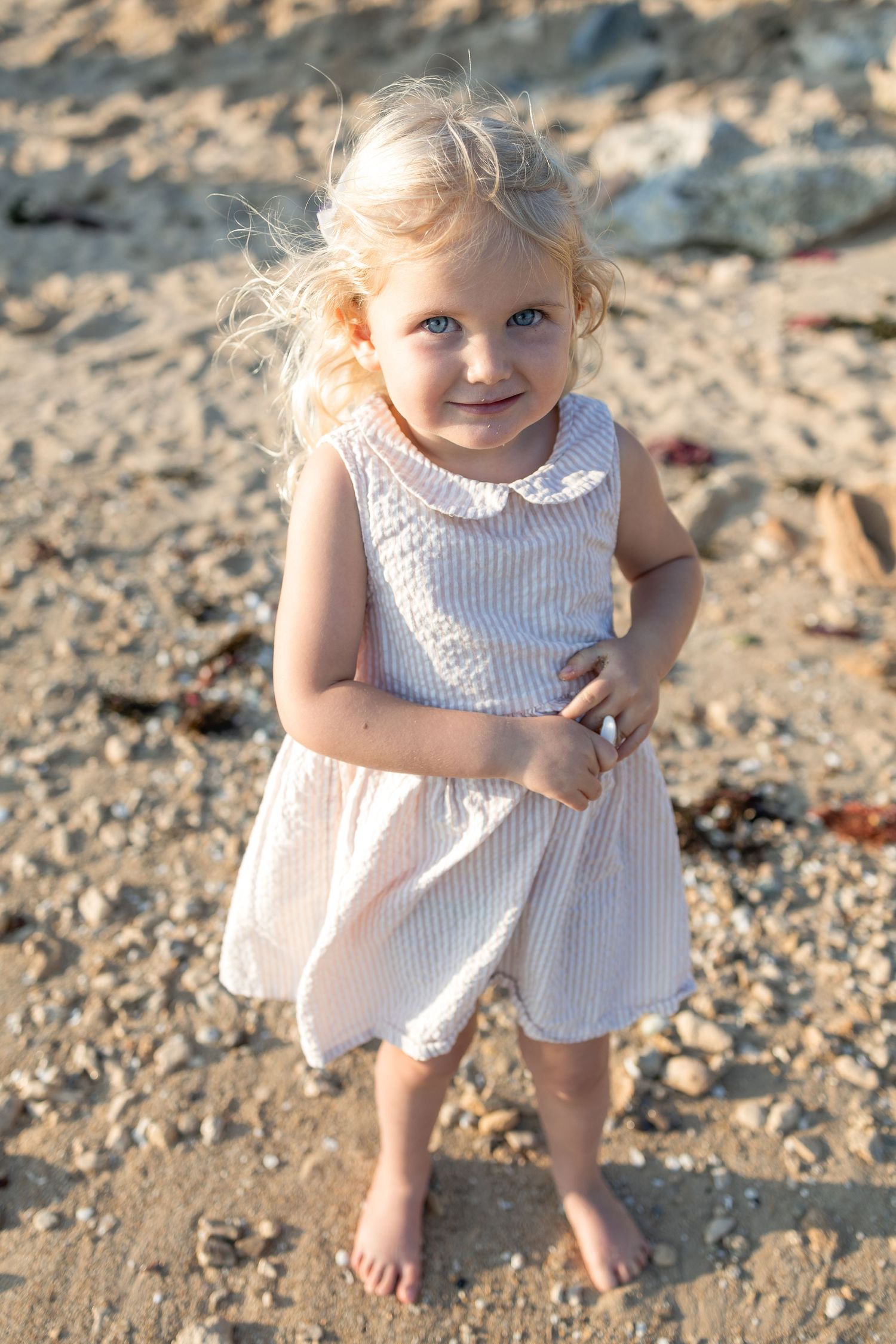 Adorable blonde toddler girl with blue eyes wearing a white dress standing barefoot on a sandy beach. Taken by Golden Hour Perth