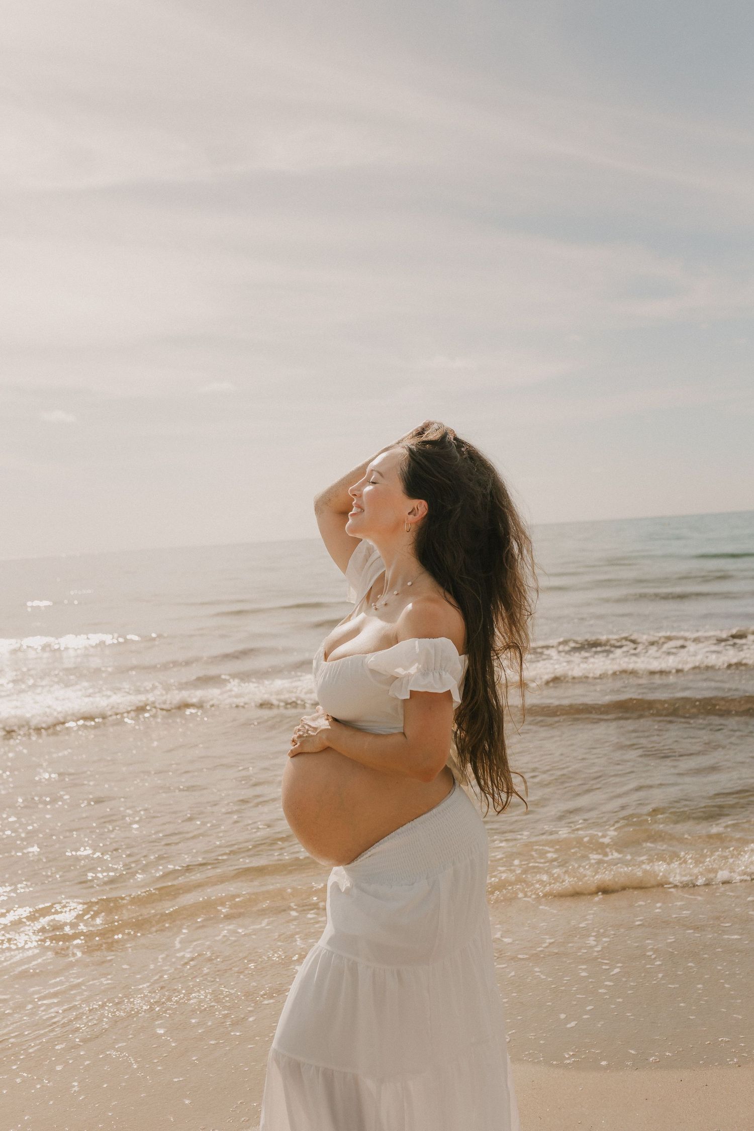 Pregnant woman in white off-shoulder dress with flowing hair cradling her bump on a sandy beach by the ocean.