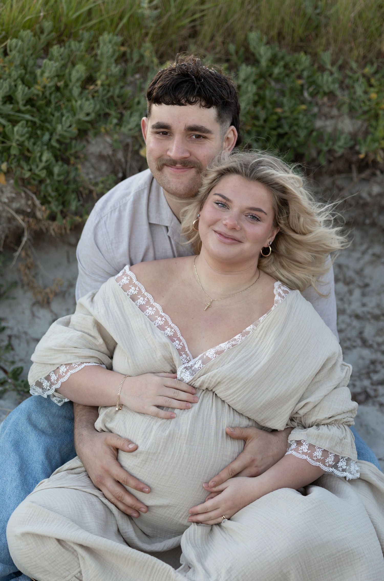 Couple poses for maternity photo on beach, man embracing pregnant woman in cream lace dress, sitting on sand near greenery.