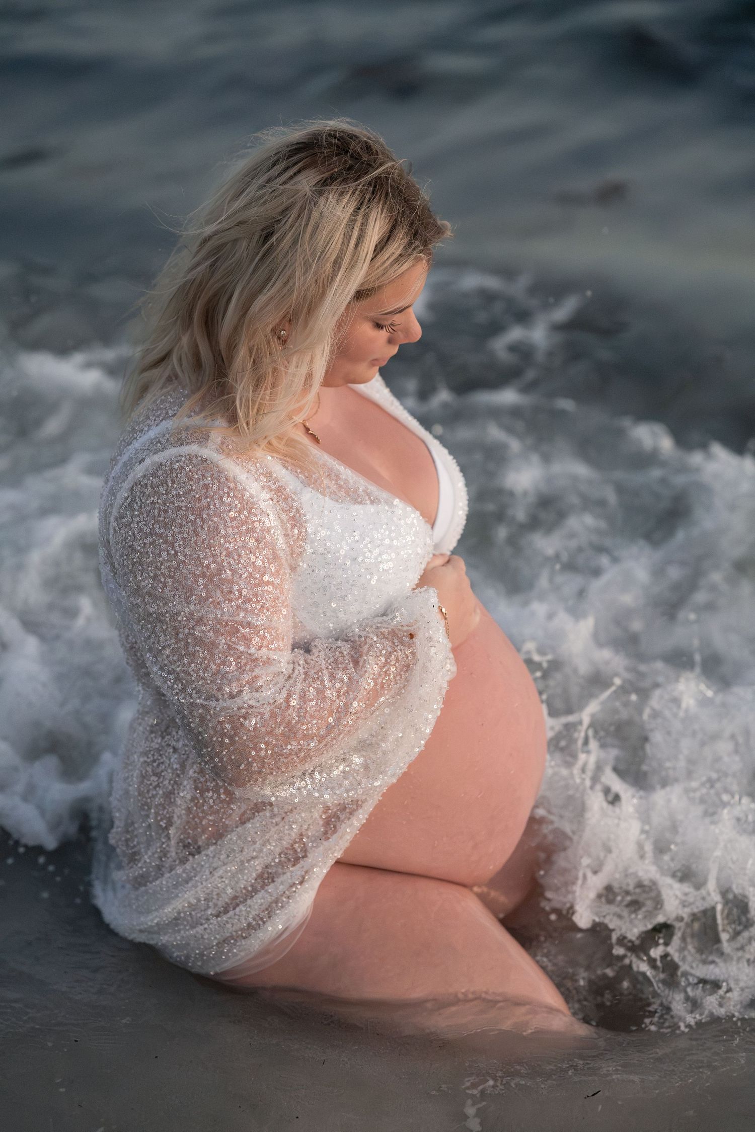 Pregnant woman in white lace robe sits in ocean waves, cradling her baby bump during a maternity photoshoot.
