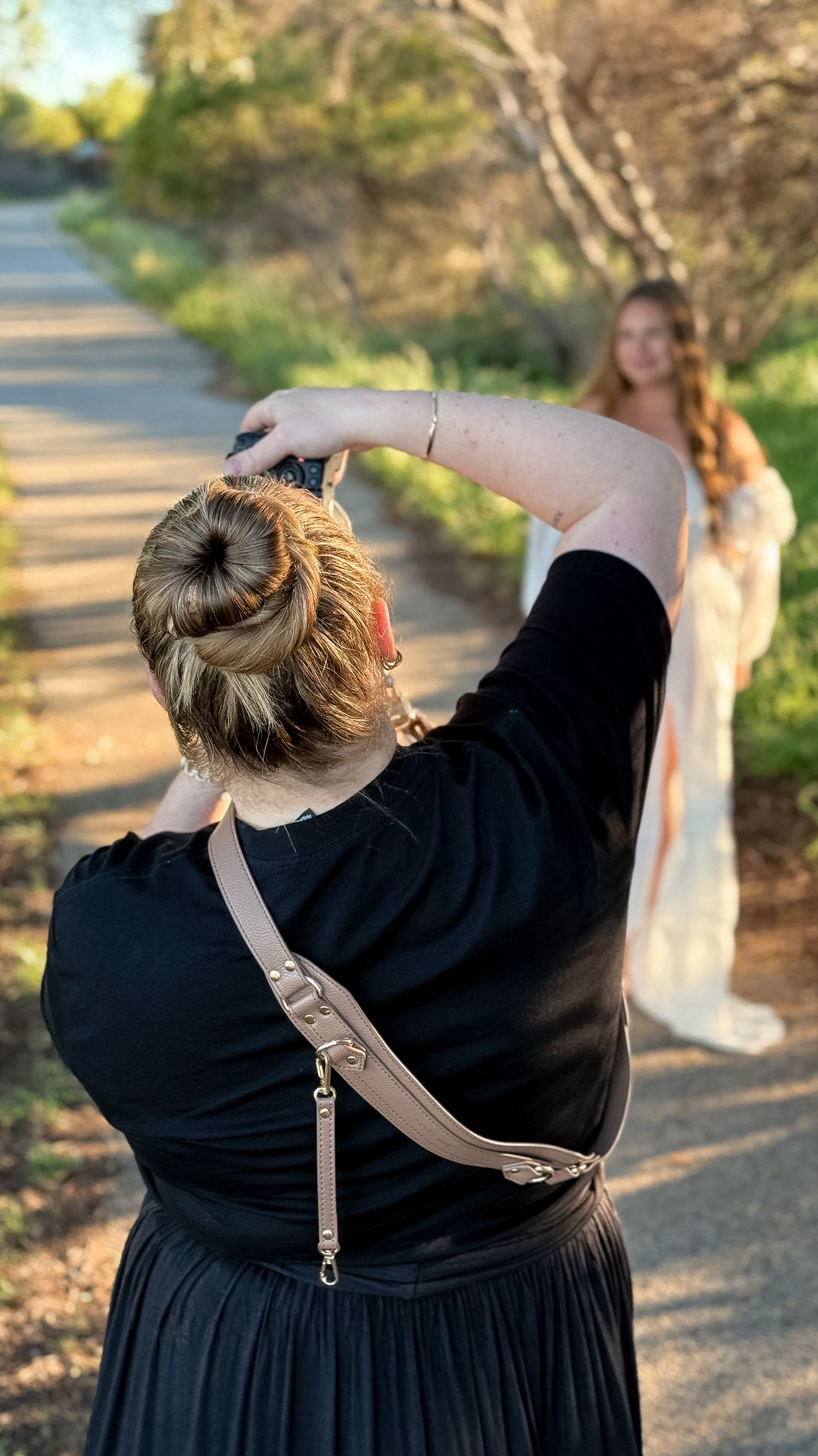 Photographer in black dress captures a woman in white gown on a sunlit tree-lined path outdoors.