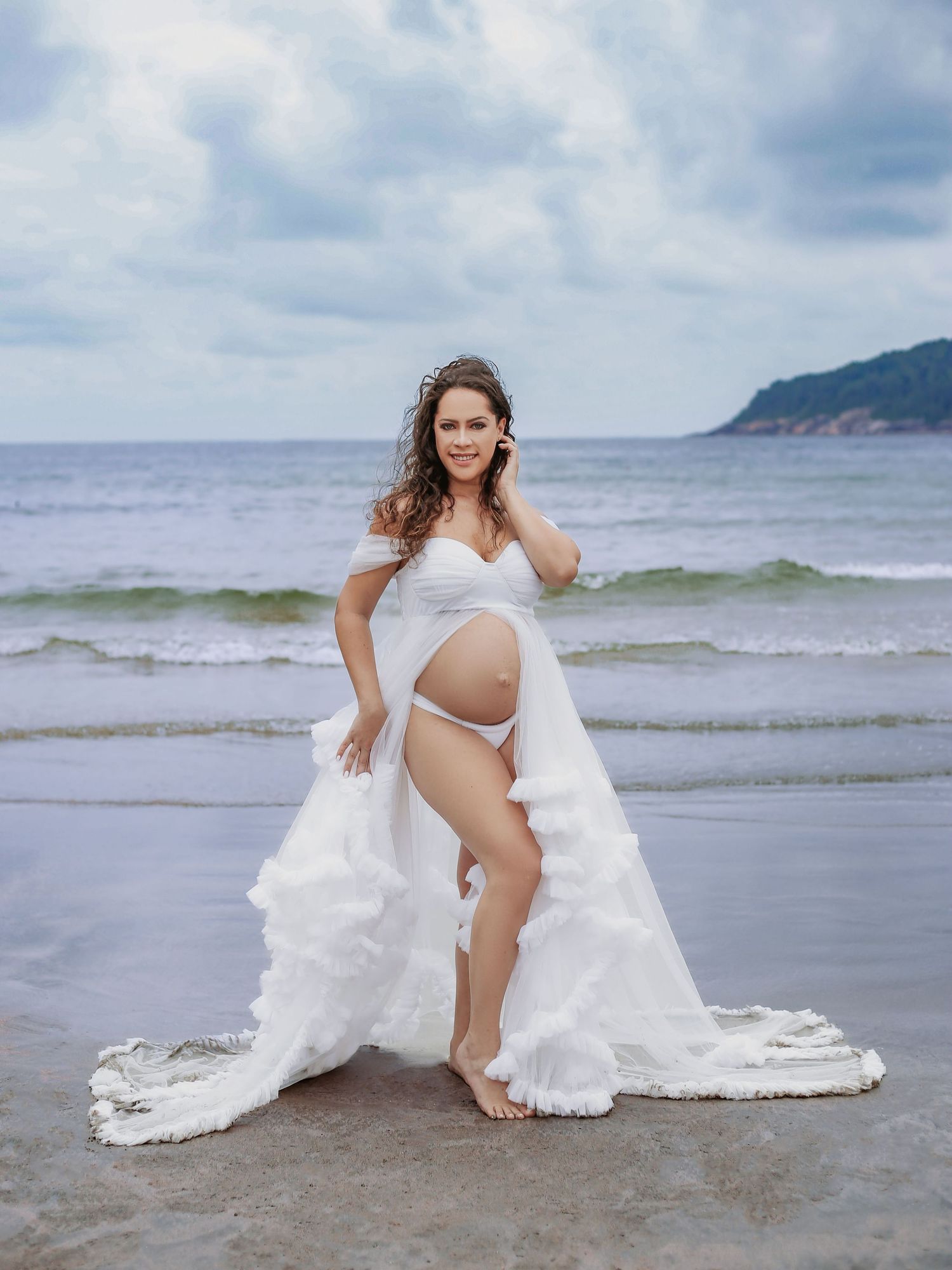 Pregnant woman in flowing white gown holding her baby bump while standing on a beach with ocean waves.