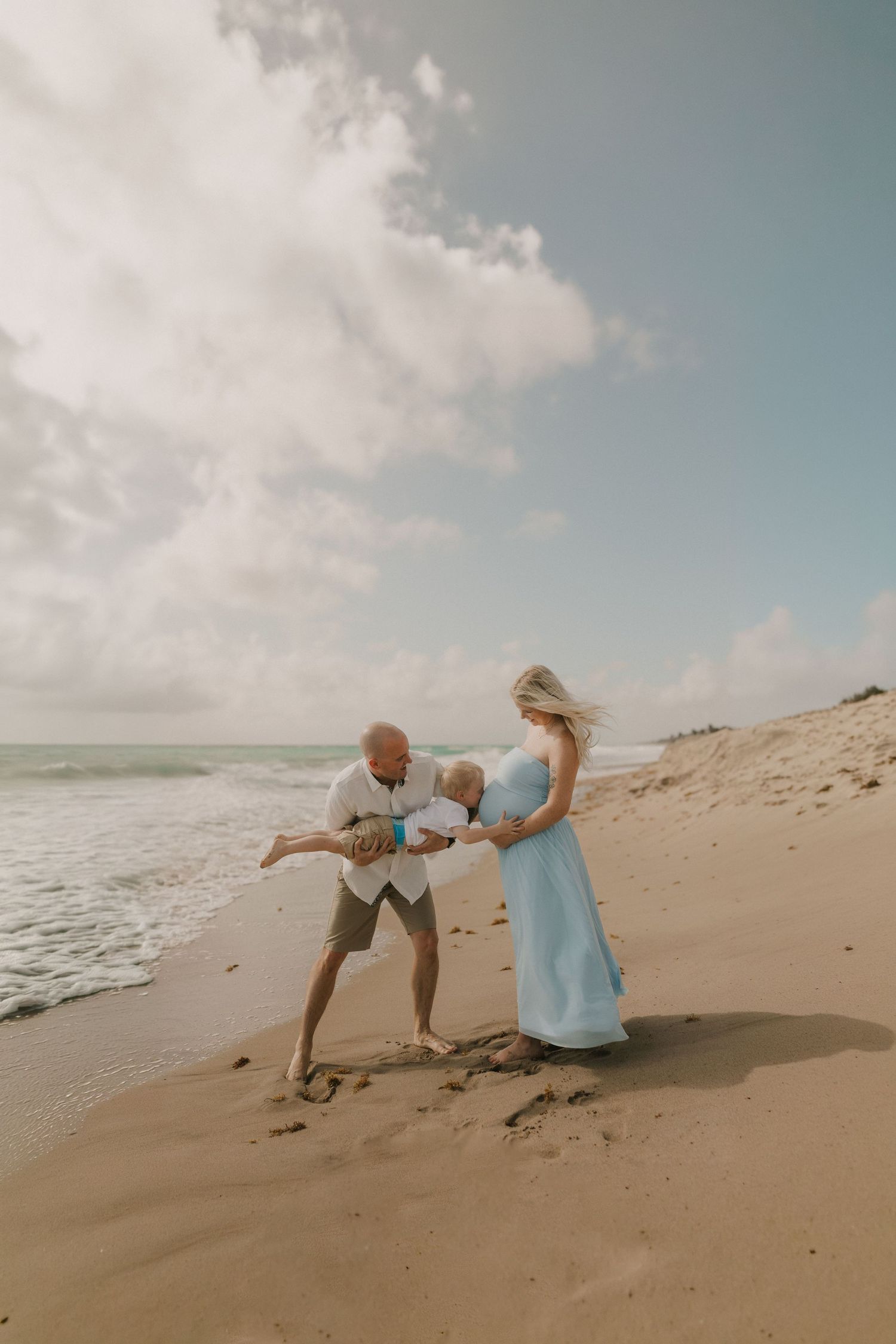 Pregnant woman in blue dress with partner and child playing on a sandy beach under a cloudy sky.