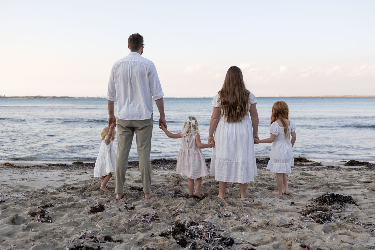 Family of four in white outfits holding hands on a sandy beach at sunset, viewed from behind. Cape Peron Perth