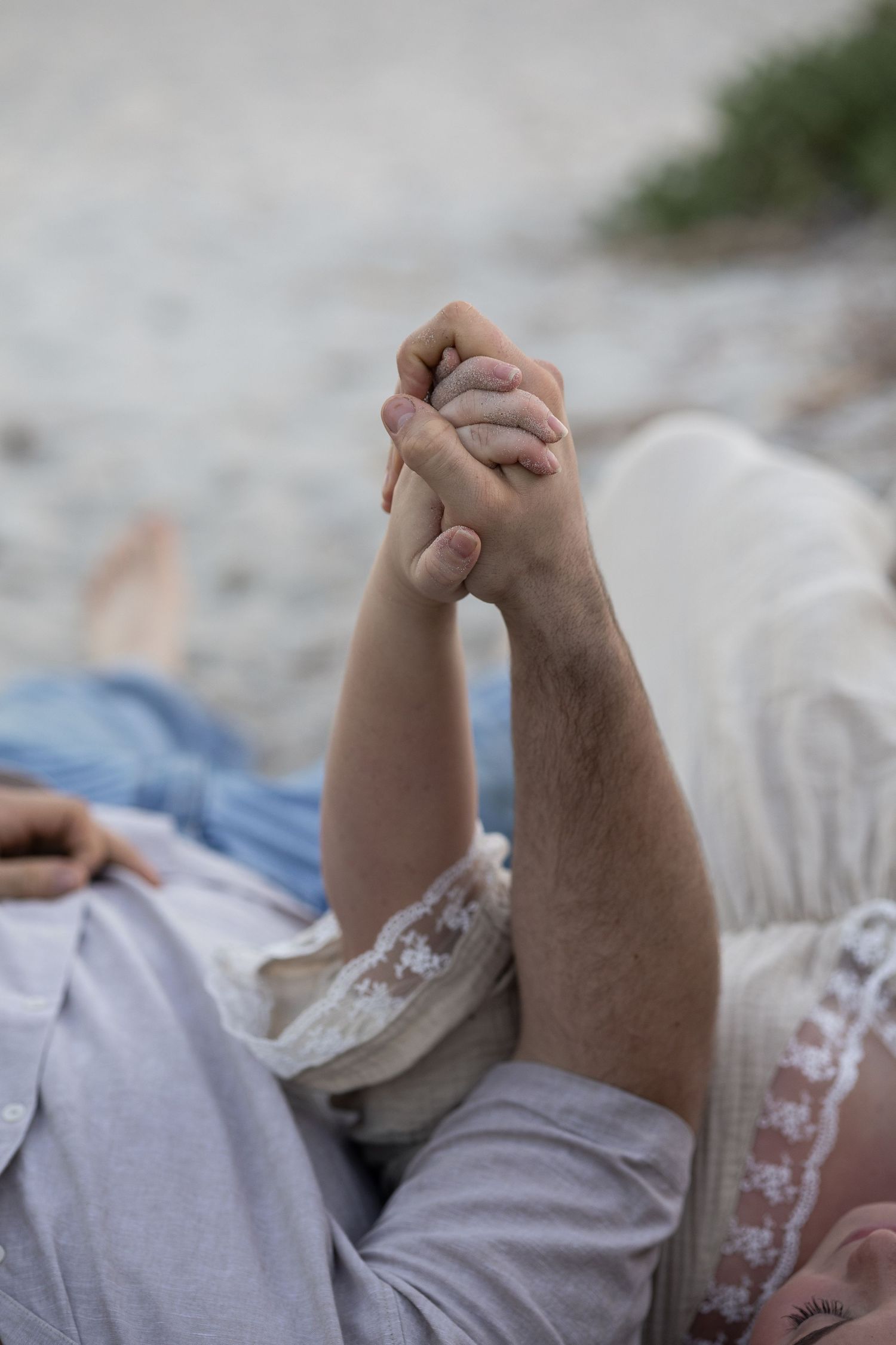 Couple holding hands while lying on a sandy beach, wearing casual light-colored clothing. Woodman Point Perth