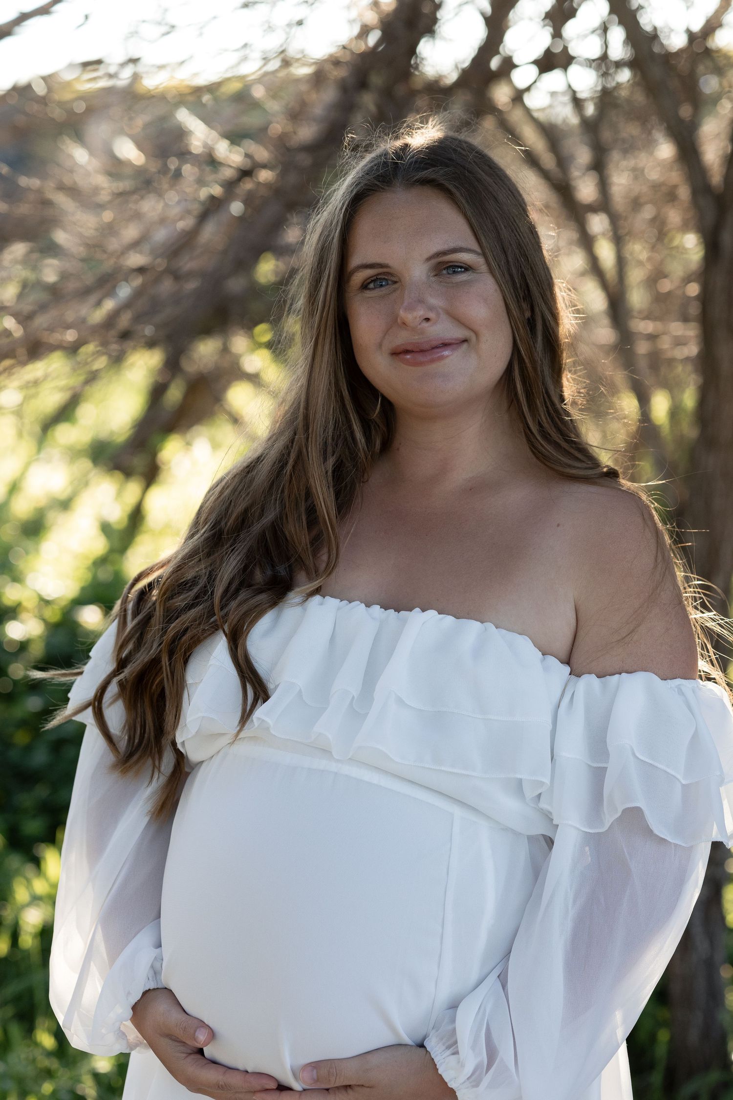 Smiling pregnant woman in white off-shoulder dress holding baby bump outdoors among trees. Woodman Point Perth