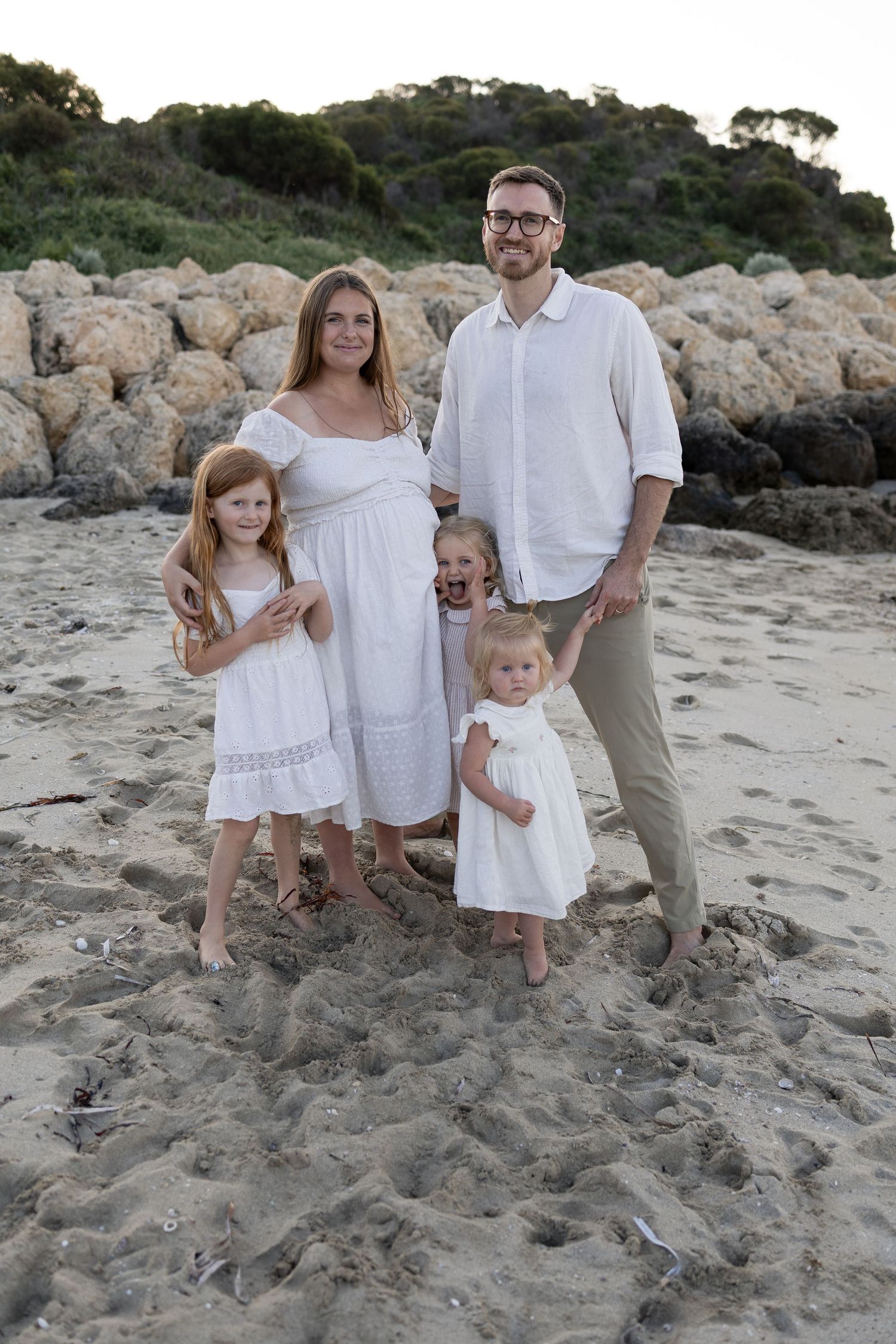 Family of five in white outfits posing on a sandy beach with rocky cliffs in the background.
