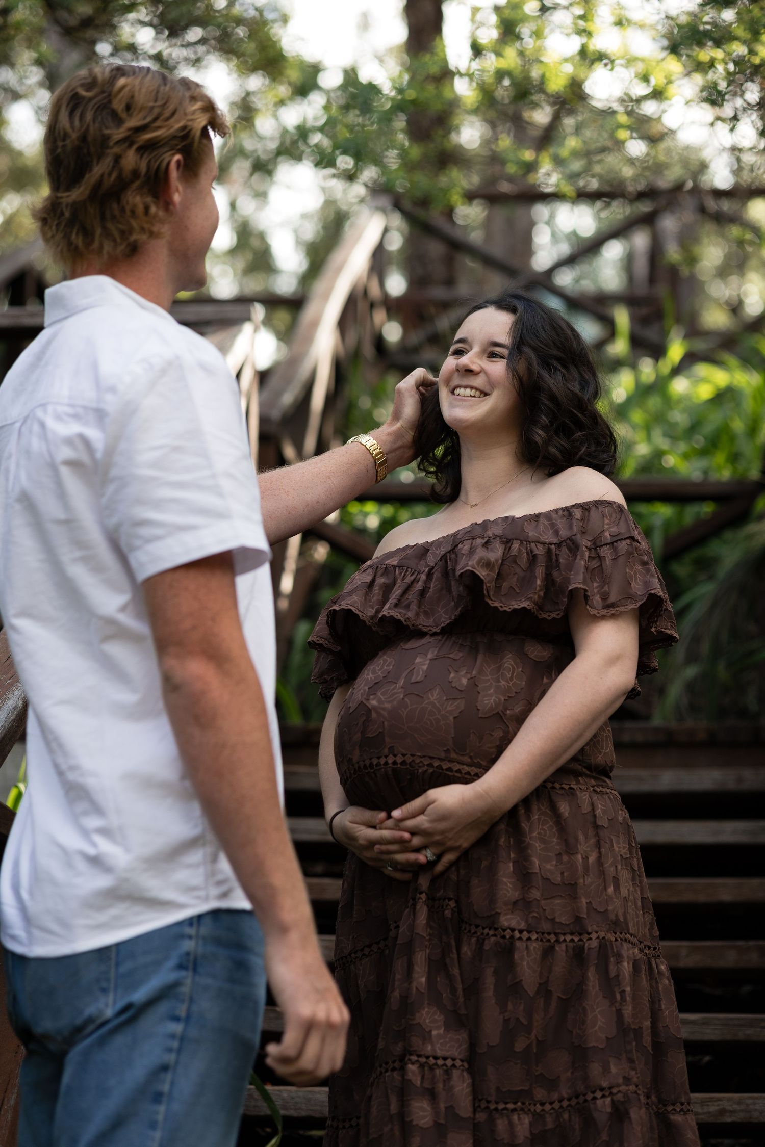 A smiling pregnant woman in a brown off-shoulder dress holds her belly as a man gently touches her face outdoors.  Araluen Perth
