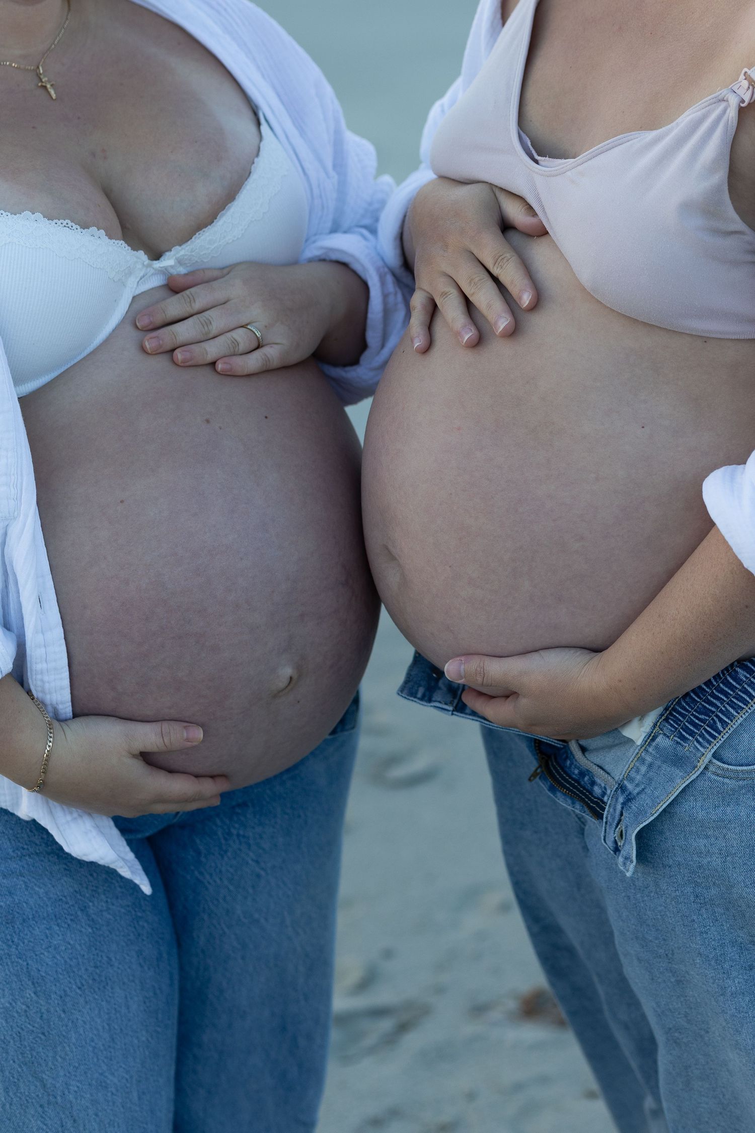 Two pregnant women in jeans and light tops standing side by side, cradling their baby bumps on a beach.