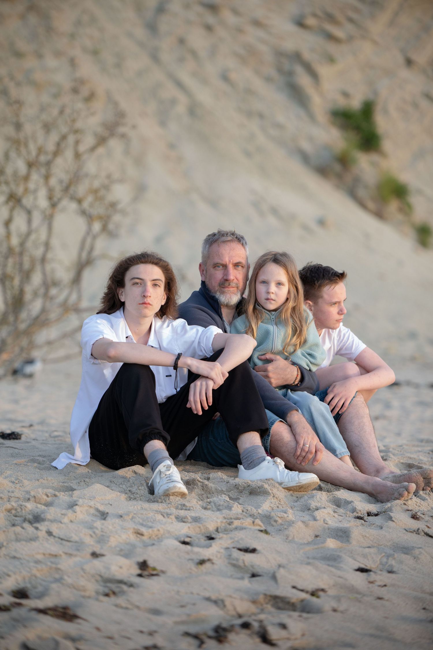 Father sitting on sandy beach with three children, posing in front of sandy dunes.