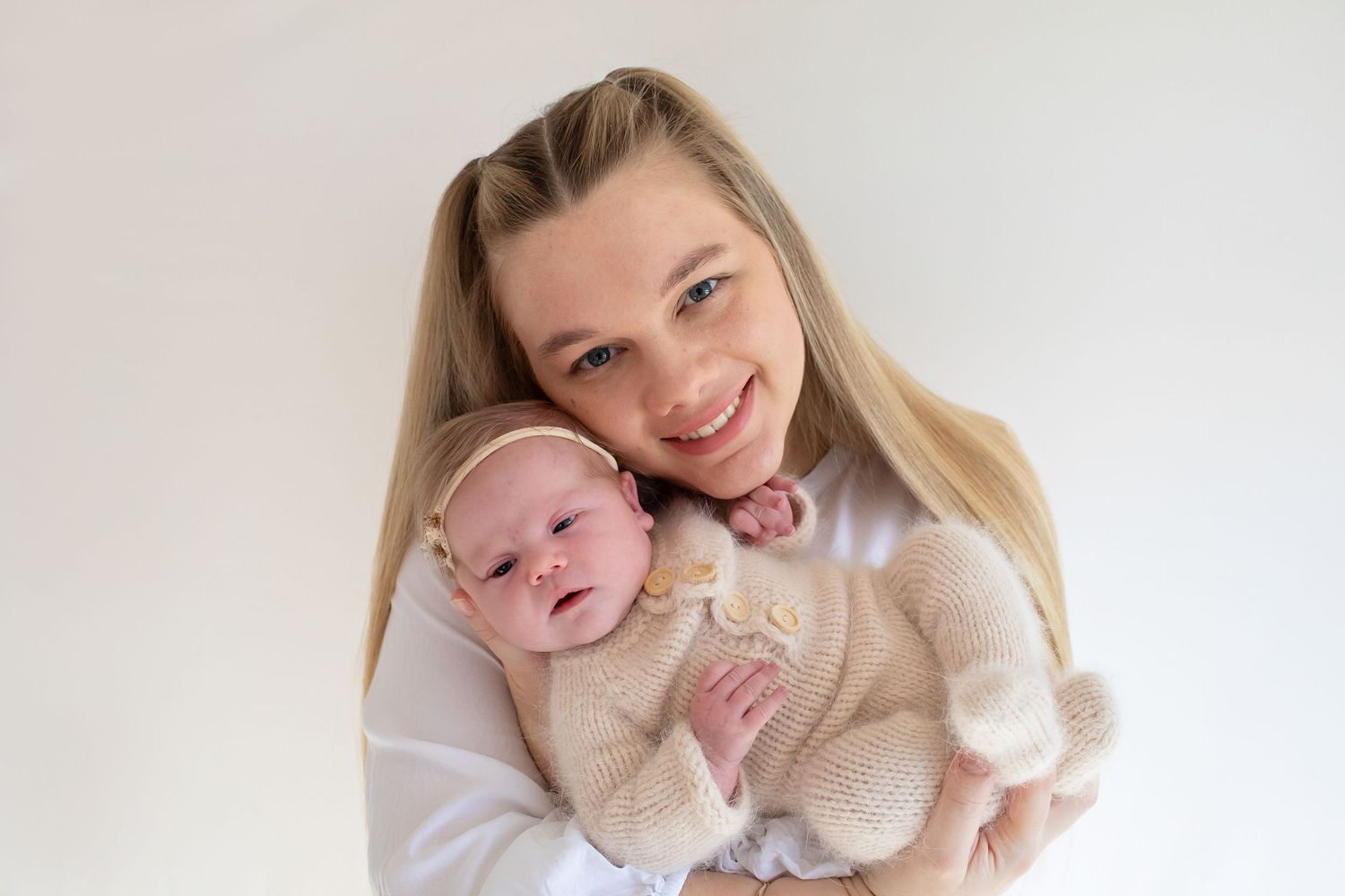 Smiling blonde mother holding her newborn baby in a cream knit outfit against white background.