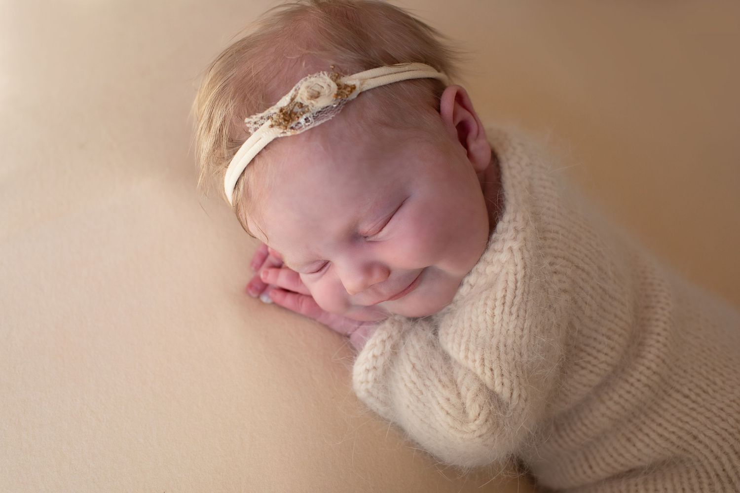 Sleeping newborn baby girl in beige knit wrap with floral headband on warm cream background.