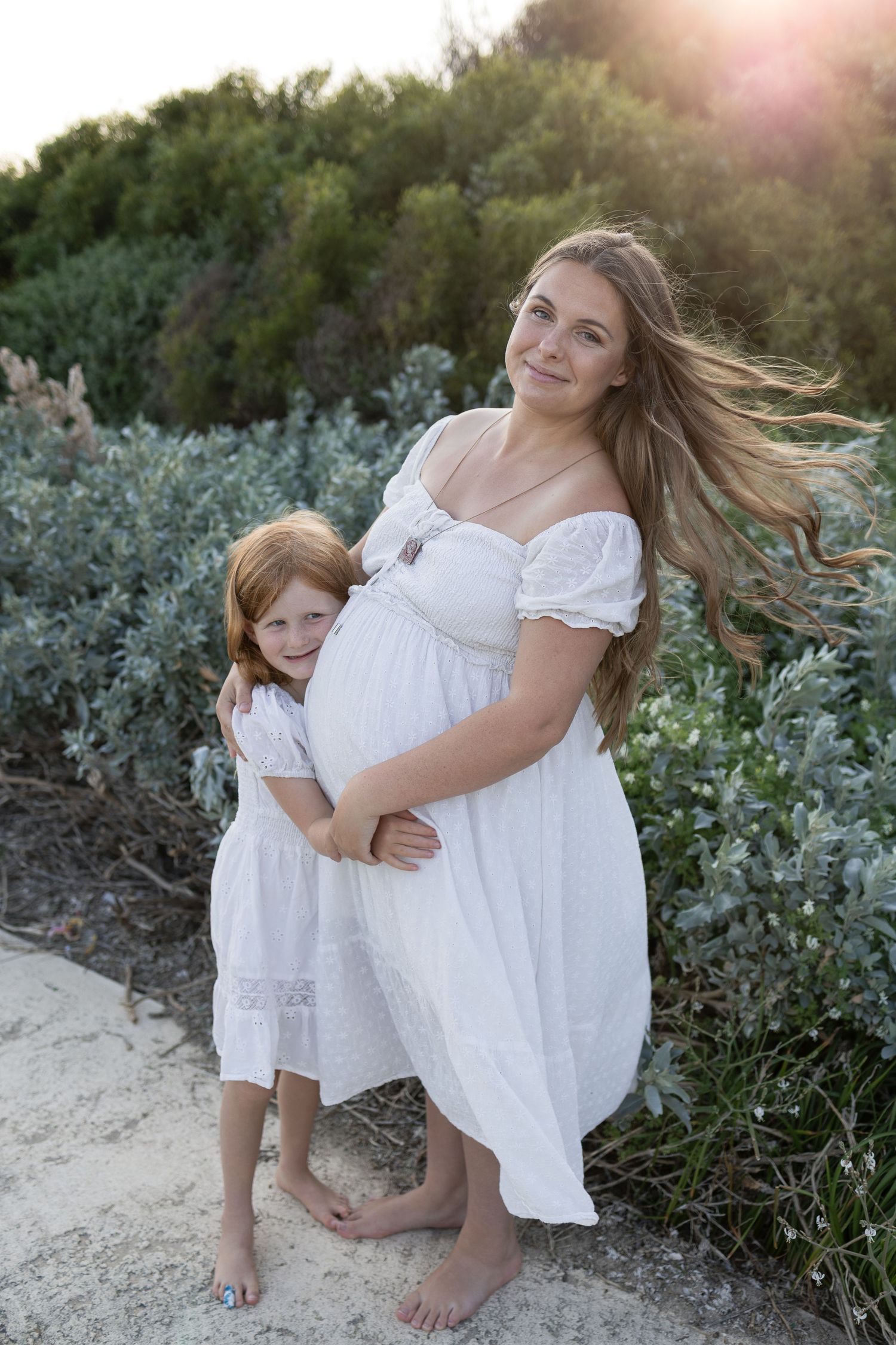 Pregnant woman in white dress with young redhead girl hugging her baby bump outdoors at golden hour. Cape Peron Perth