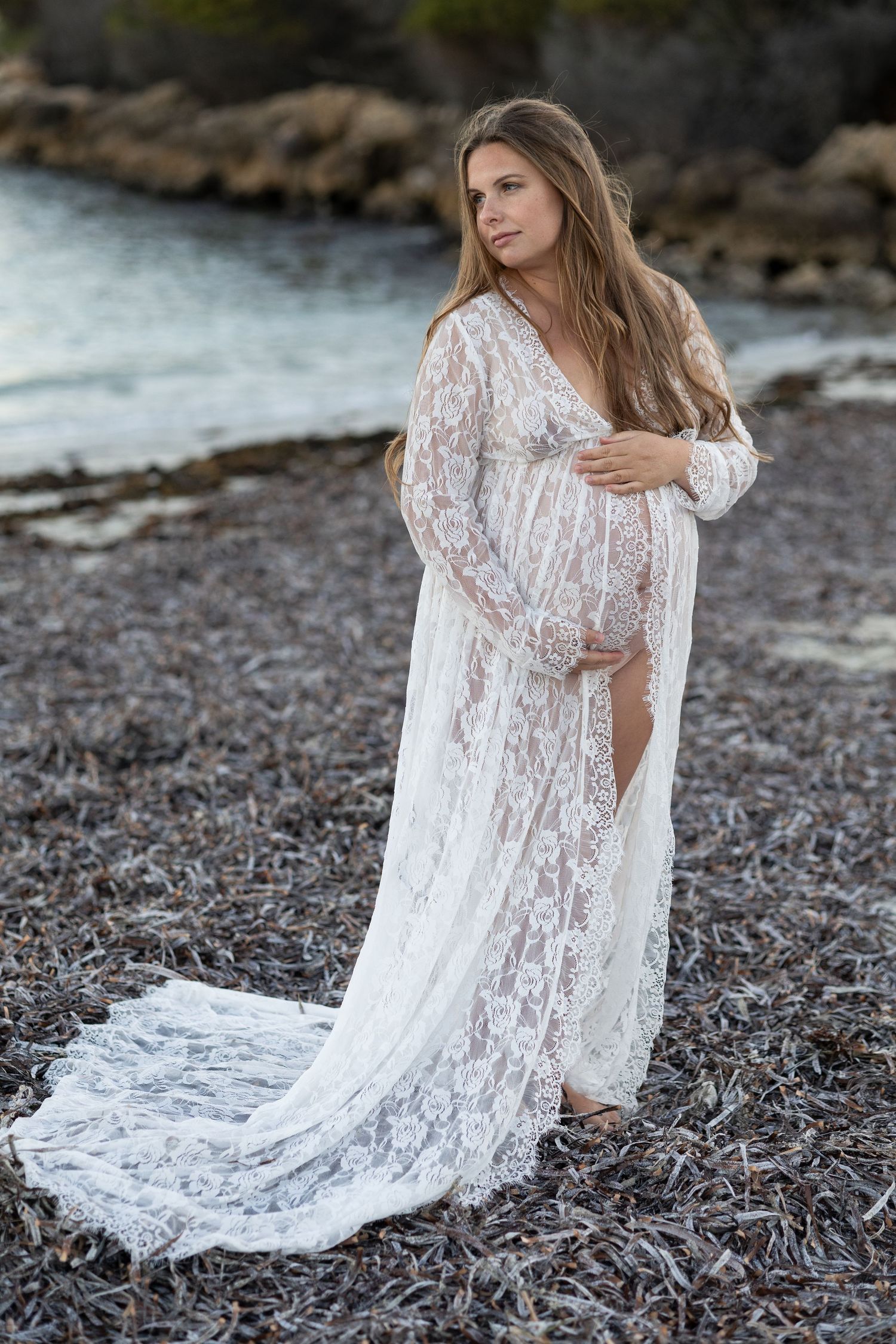 Pregnant woman in elegant white lace gown posing on rocky shoreline near water during maternity photoshoot.
