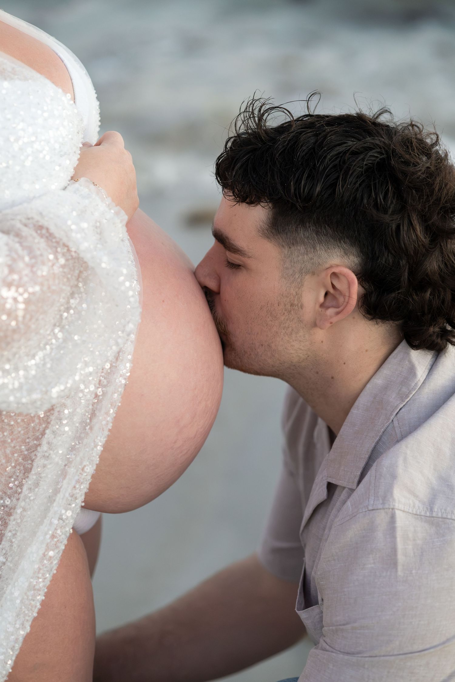 Man tenderly kisses pregnant woman's baby bump during a romantic beach maternity photoshoot.