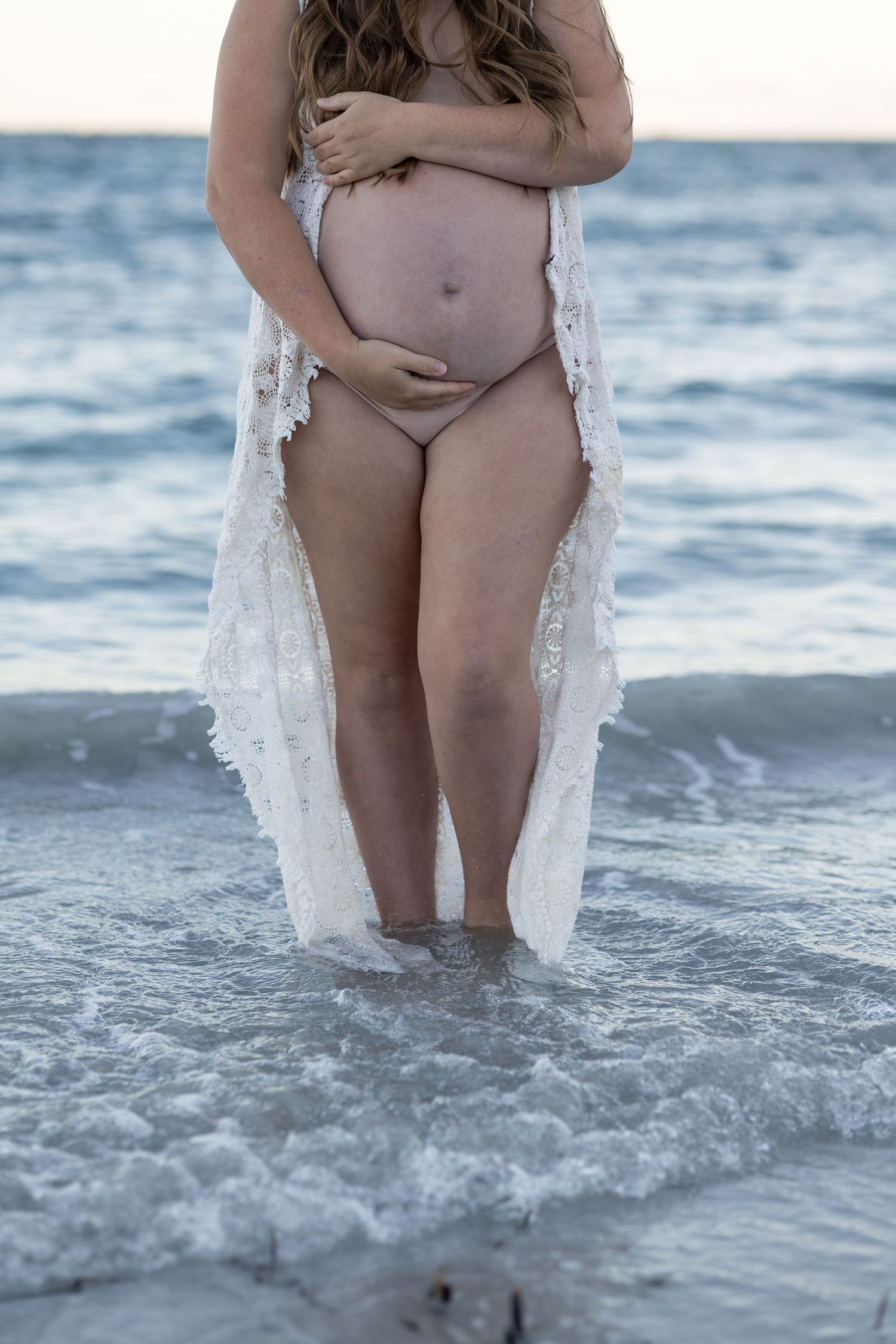 Pregnant woman standing in ocean waves wearing white lace robe, cradling bare bump.