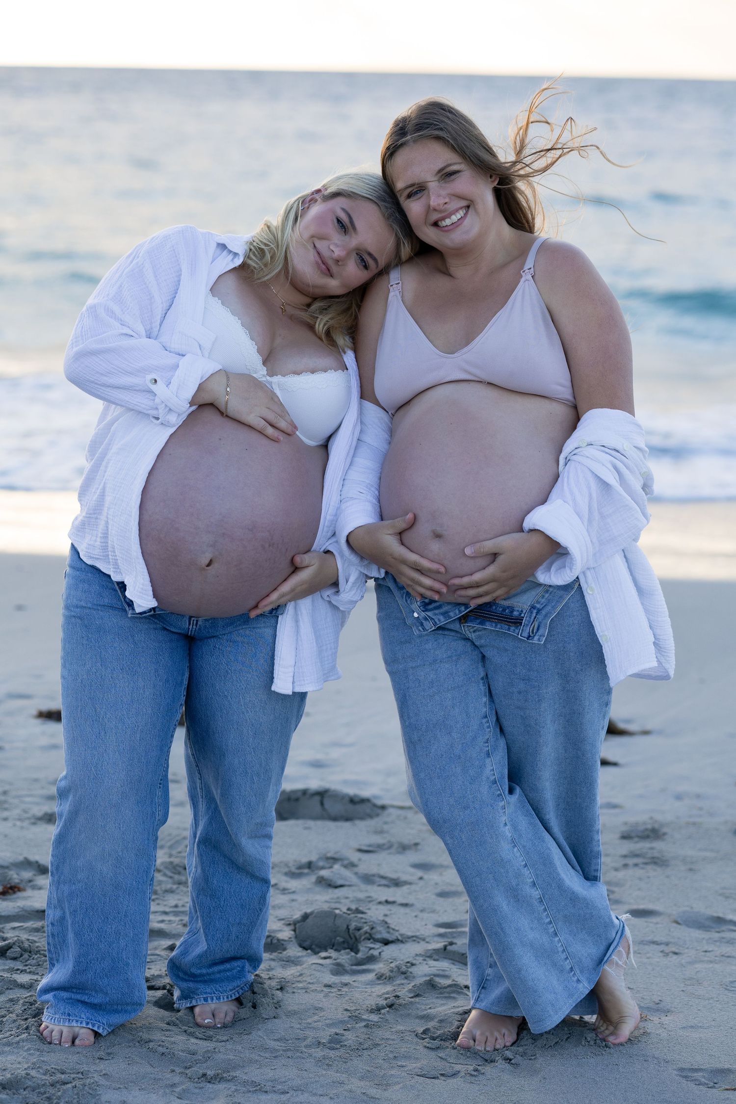 Two pregnant women smiling on a beach, wearing jeans and crop tops, showing their baby bumps at sunset.