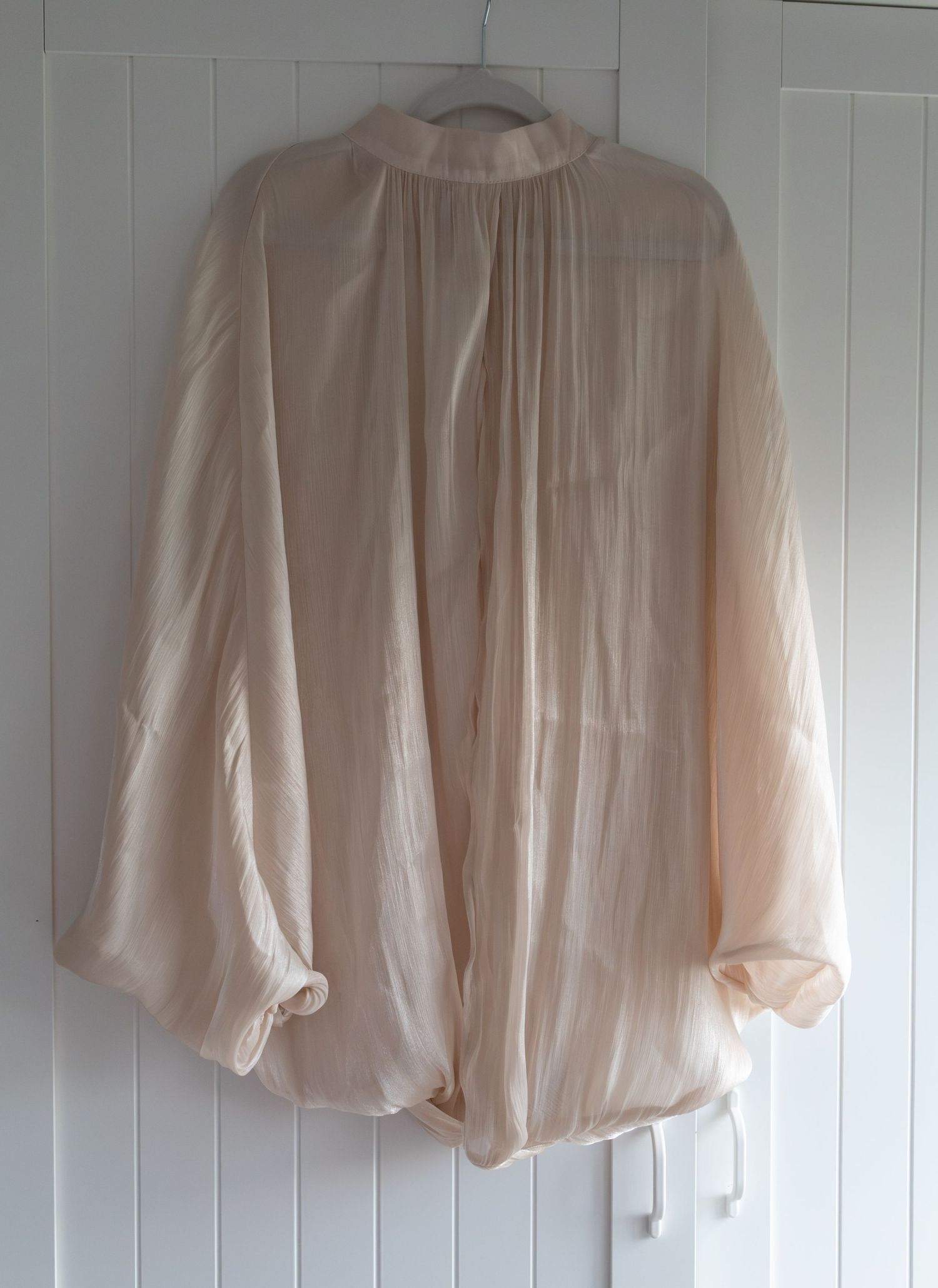 Sheer blush pink oversized blouse with balloon sleeves, gathered neckline, and tie hem, hanging on a white door.