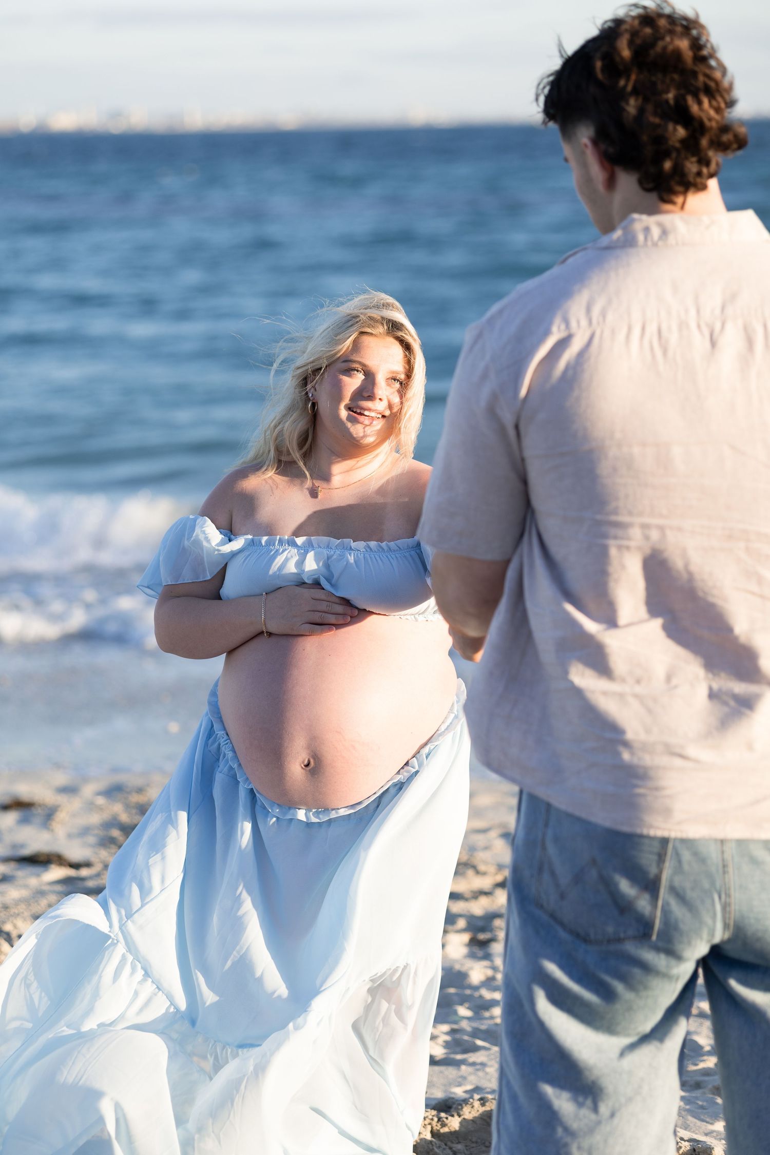 Smiling pregnant woman in blue off-shoulder dress holds her bare baby bump while facing her partner on a sunny beach. Woodman Point Perth