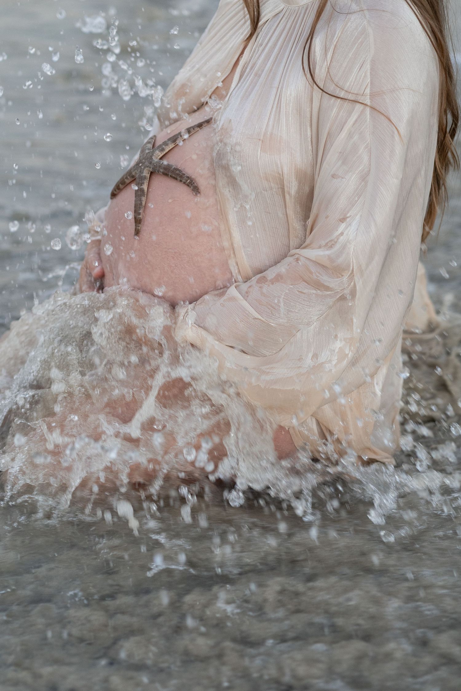 Pregnant woman in wet sheer dress standing in ocean waves with a starfish on her baby bump. Woodman Point Perth