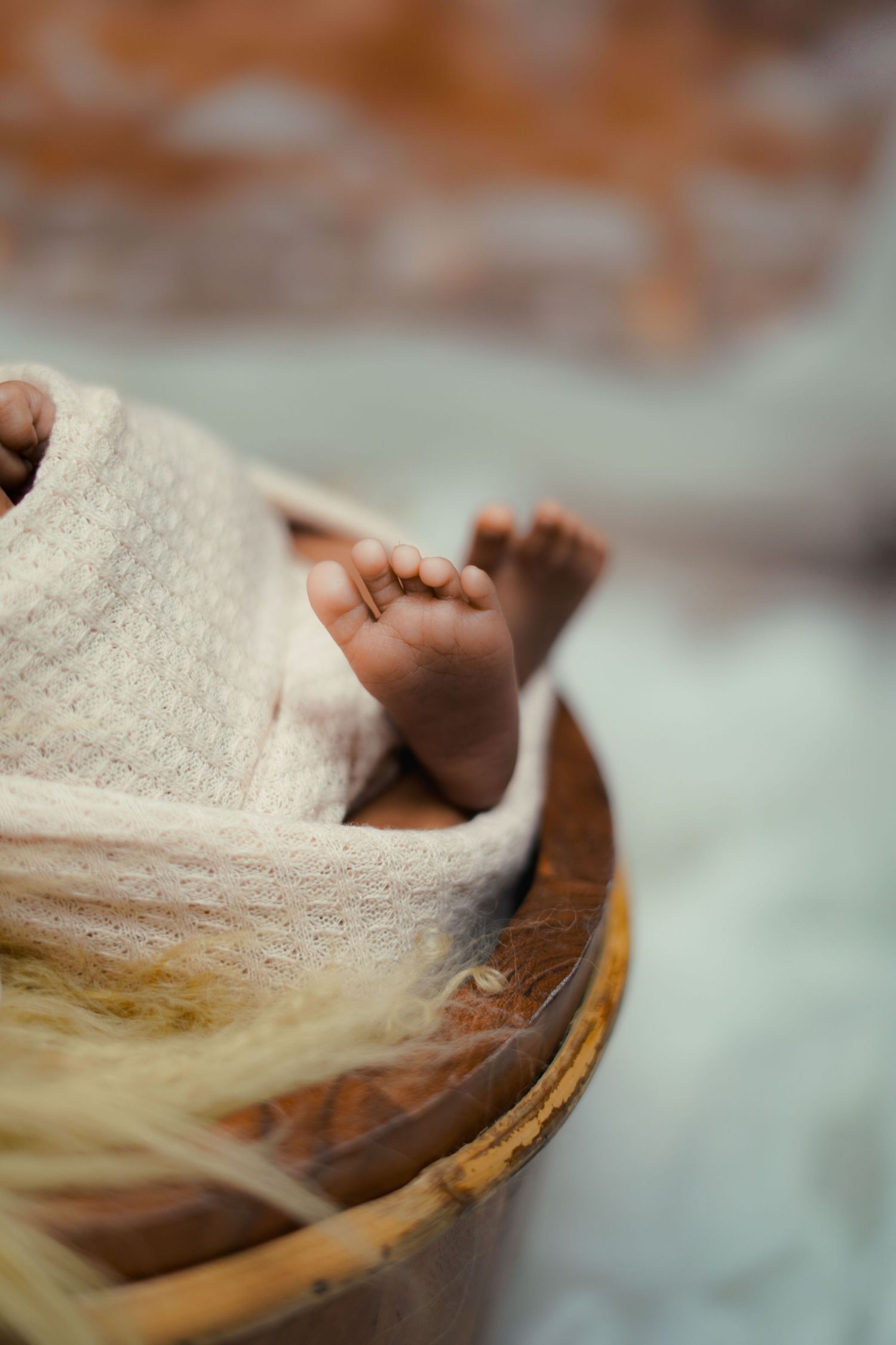 Close-up of newborn baby feet peeking out from a white knit wrap inside a wooden bowl with straw.