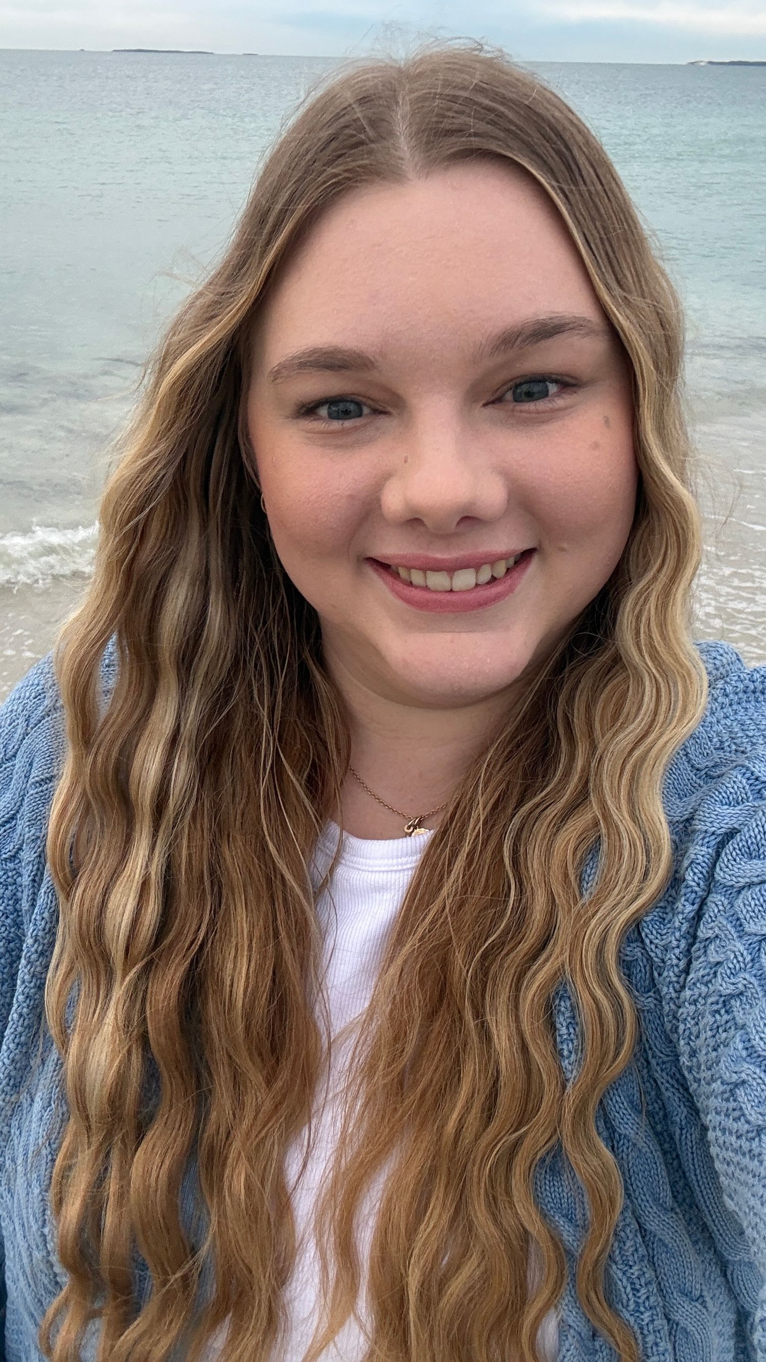Smiling teenage girl with long wavy blonde hair in blue cardigan posing at the ocean shore.
