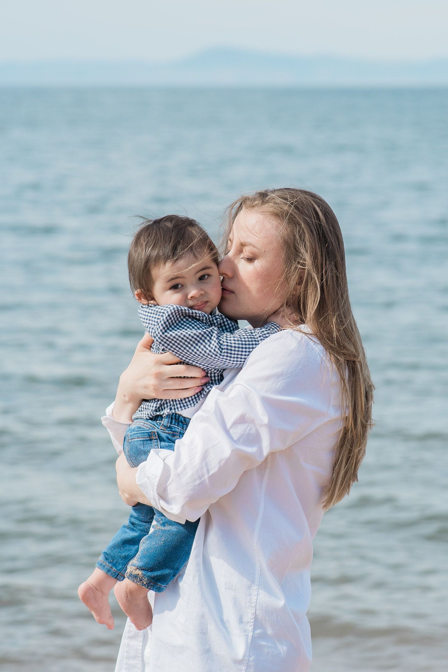 Loving mother in white shirt kissing her toddler son on the cheek by the ocean shore.
