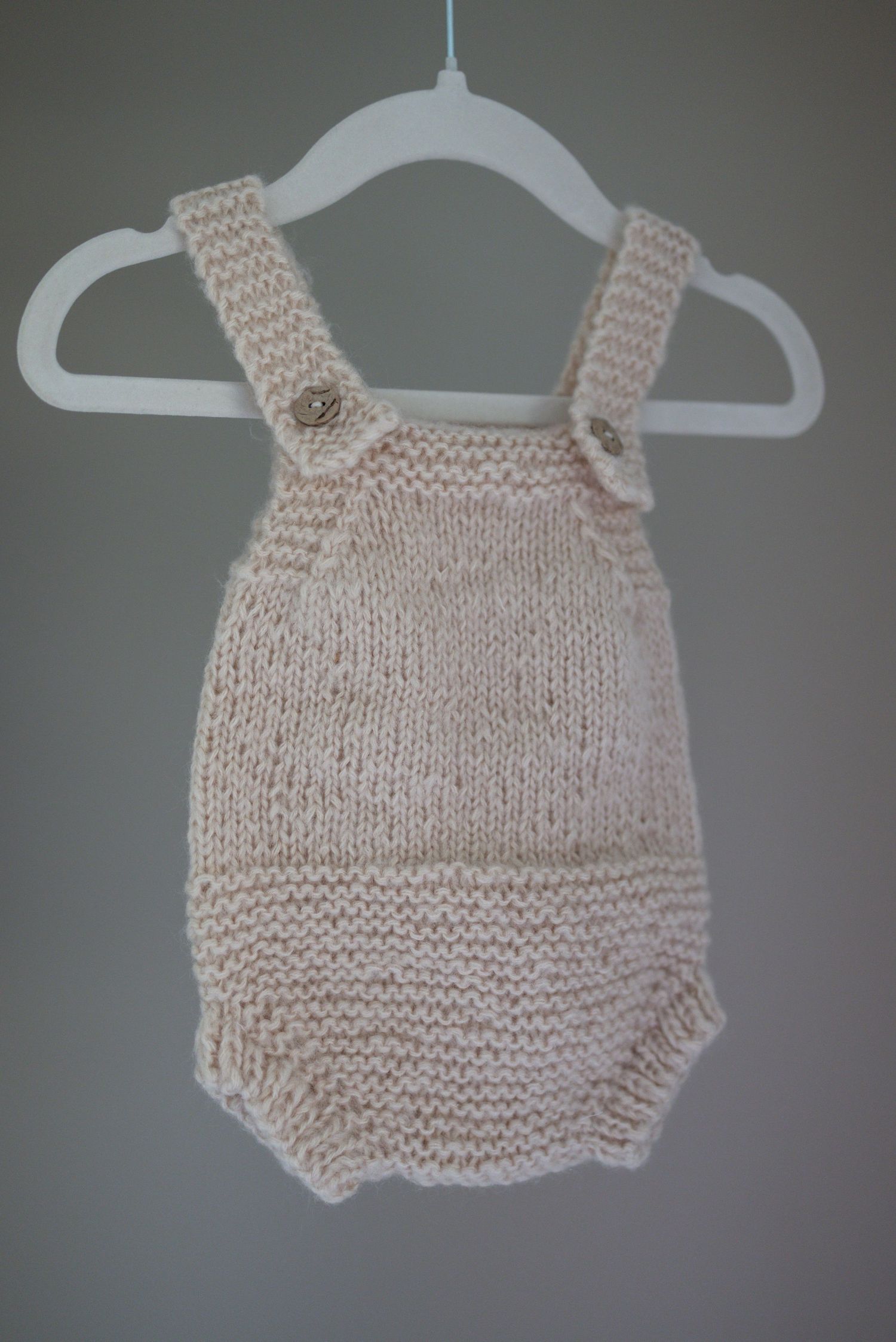 Hand-knitted cream baby romper with button straps displayed on a white hanger against a gray background. Taken by Golden Hour Perth