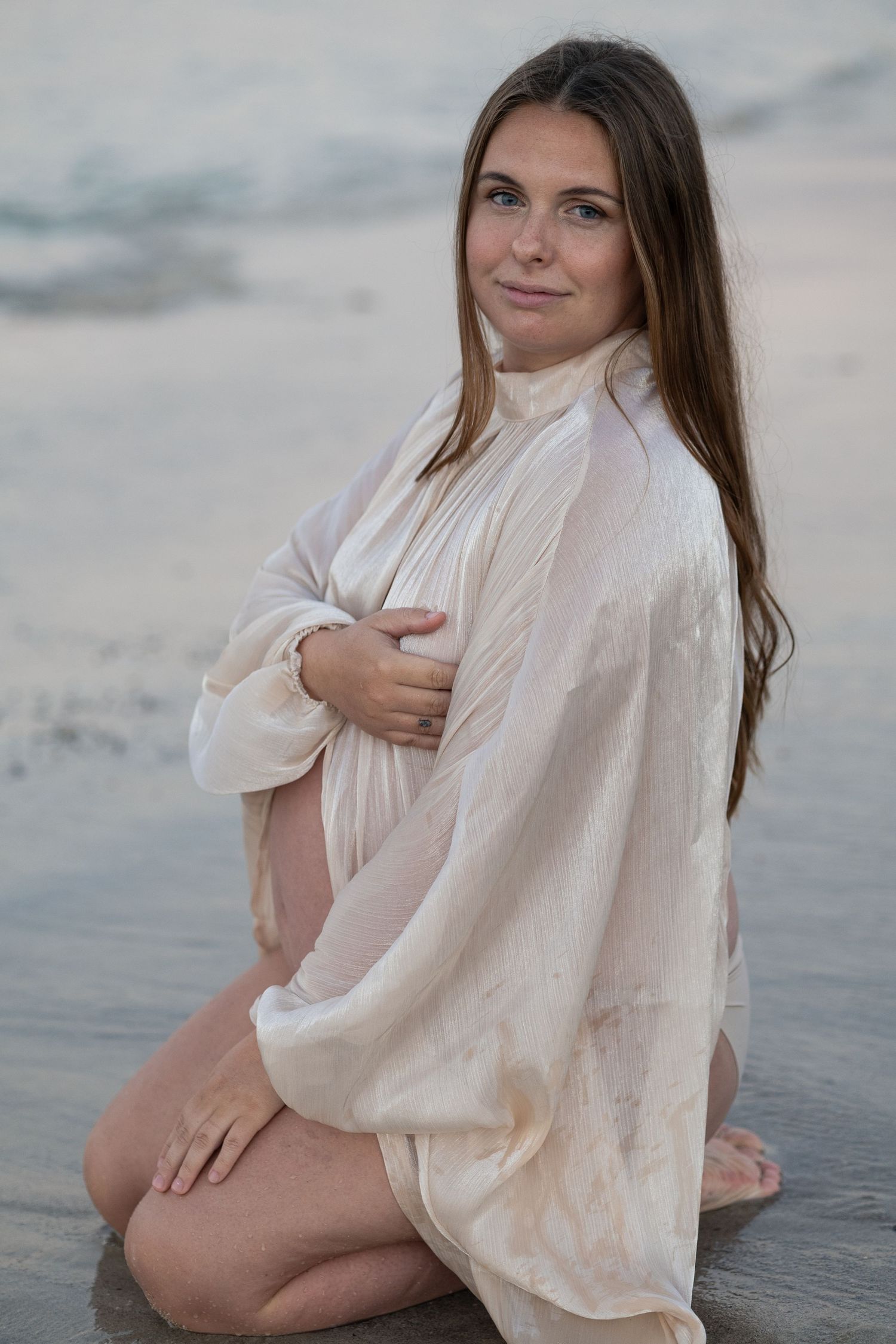 Pregnant woman in flowing white sheer top kneeling on sandy beach by ocean waves, cradling her baby bump.