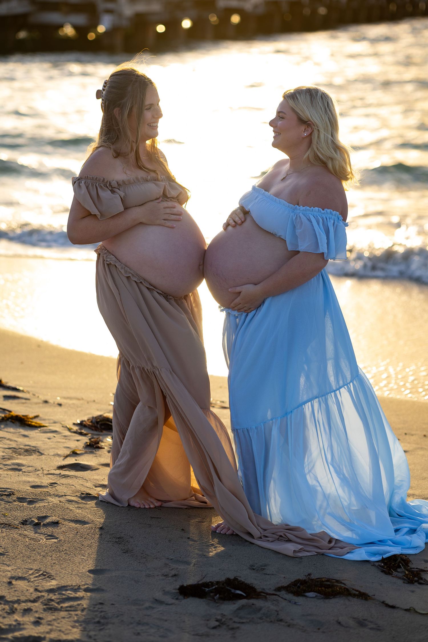 Two pregnant women in elegant maternity gowns pose together on a sunlit beach, cradling their baby bumps.