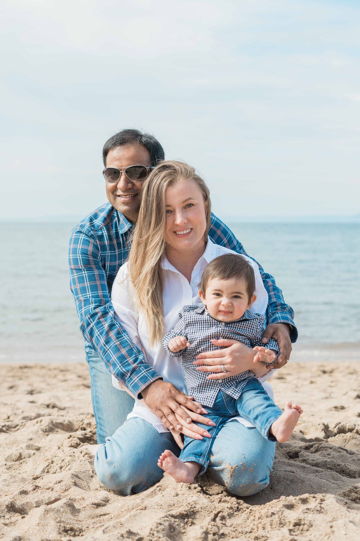 Smiling mixed-race family with baby boy posing together on a sunny beach by the ocean.