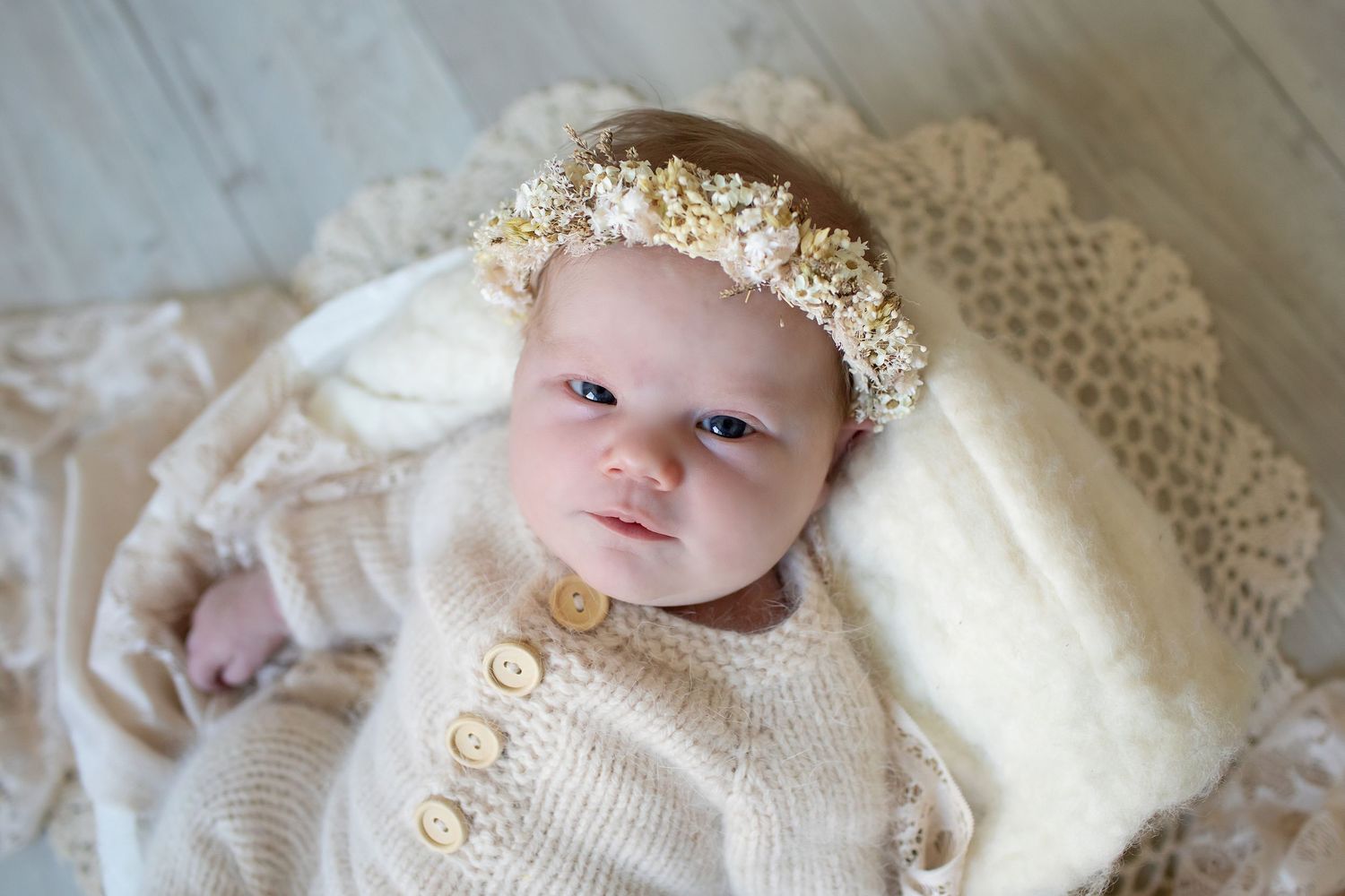 Newborn baby in beige knit outfit and floral headband lying on cream lace blanket during professional photo session.