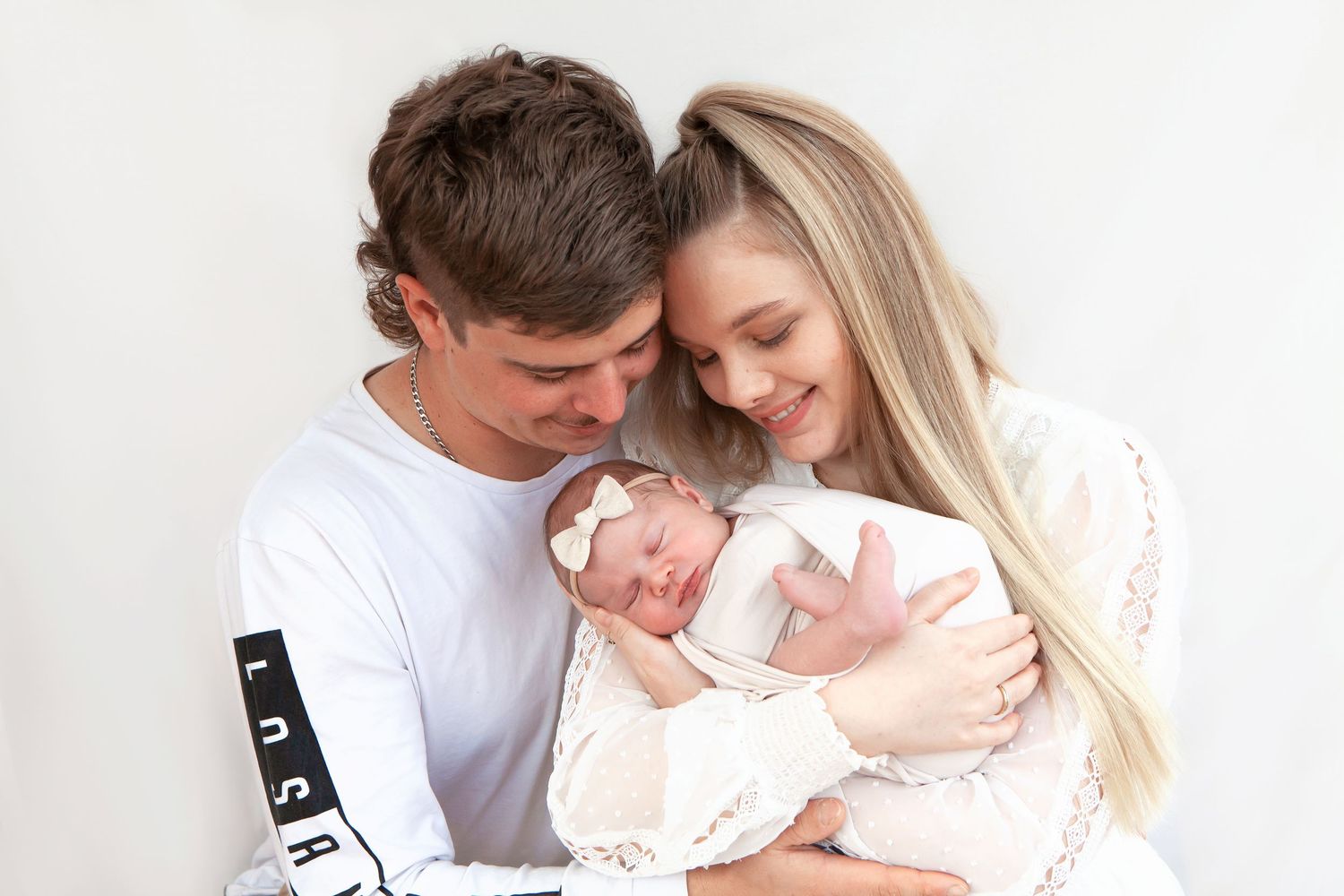 Young couple tenderly gazing at their newborn baby wrapped in white lace against a white background.