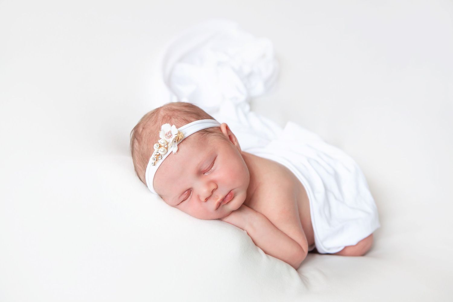 Peaceful newborn baby sleeping wrapped in white fabric with floral headband on white background.