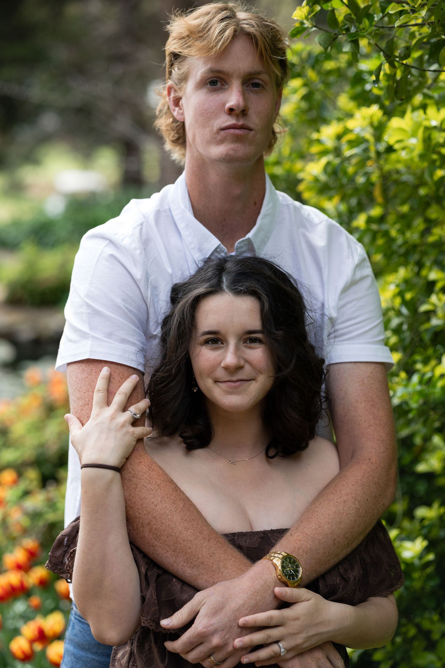 Young couple posing outdoors in a garden with orange flowers, man in white shirt embracing woman with dark hair.  Araluen Perth