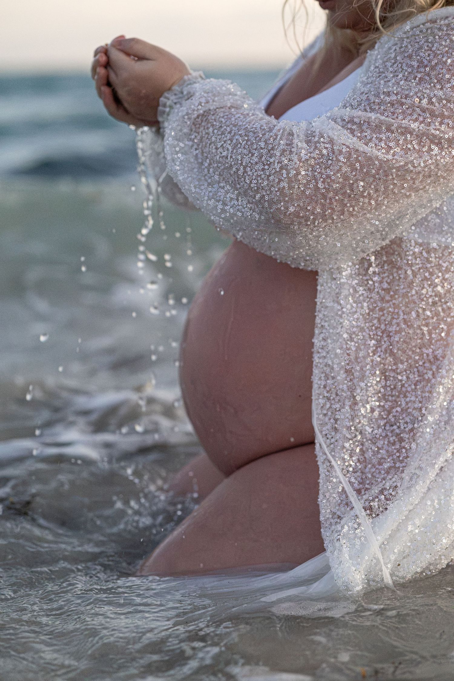 Pregnant woman in sheer white cover-up kneeling in ocean waves, letting water pour through her hands. Woodman Point Perth