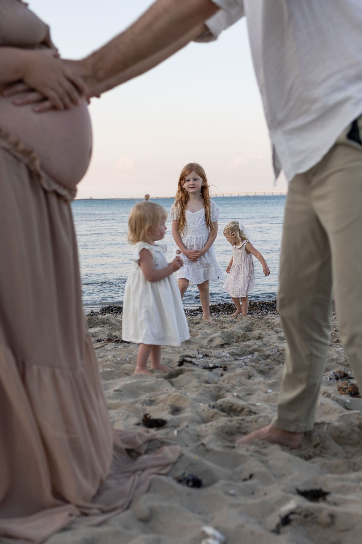 Pregnant woman and man frame three young girls in white dresses playing on a sandy beach at sunset. Taken by Golden Hour Perth