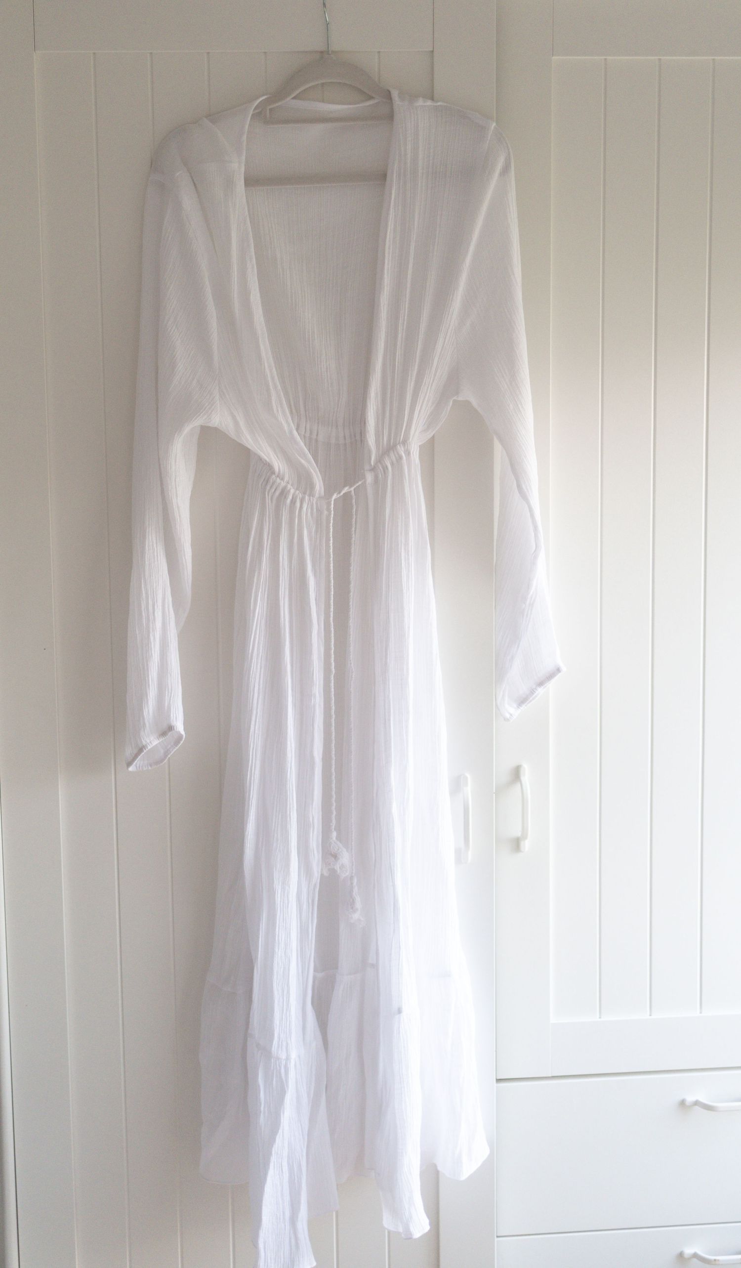White long sleeve maxi dress with deep V-neck, cinched waist, and tiered skirt hanging on a white wall.