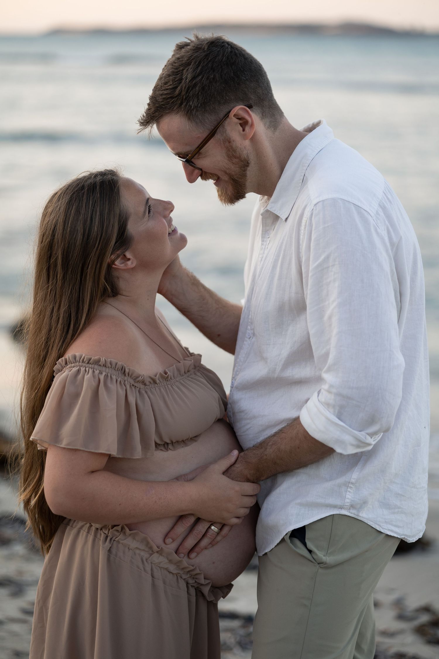 Couple sharing a tender moment on the beach, man in white shirt embracing pregnant woman in tan off-shoulder dress.