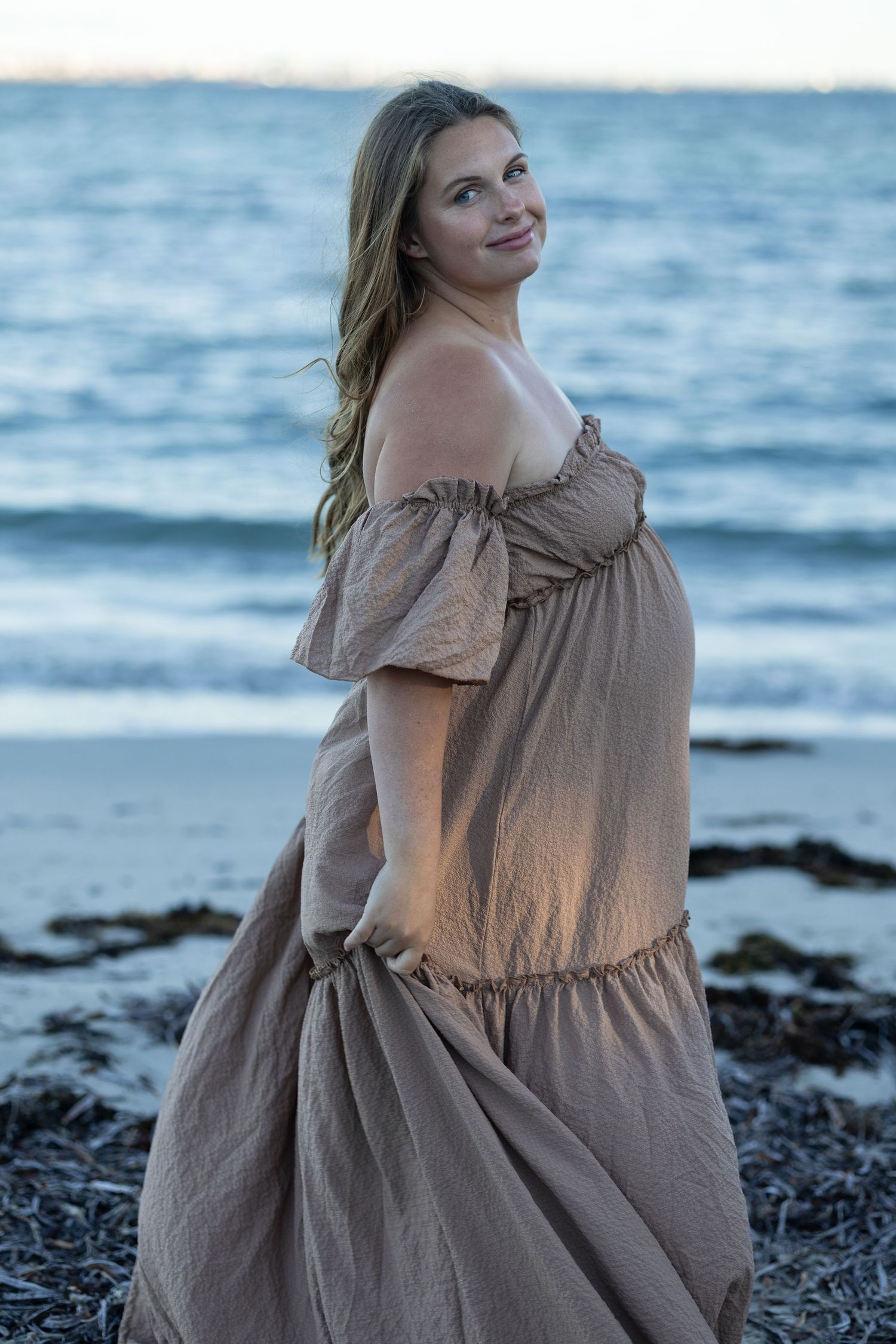 Pregnant woman in a flowing off-shoulder beige dress standing on a beach at dusk. Woodman Point Perth