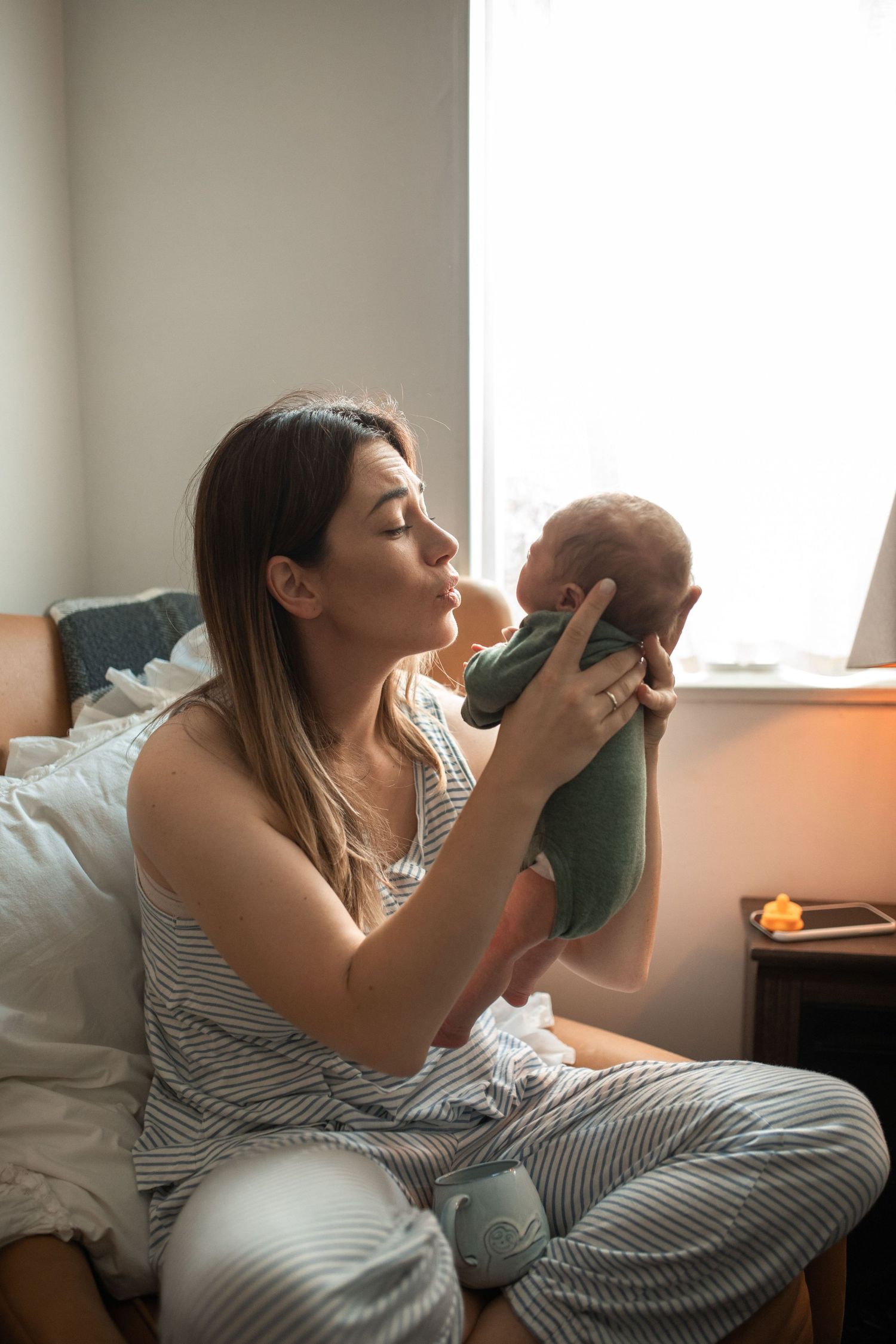 Young mother sitting on bed kissing her newborn baby near a bright sunlit window.