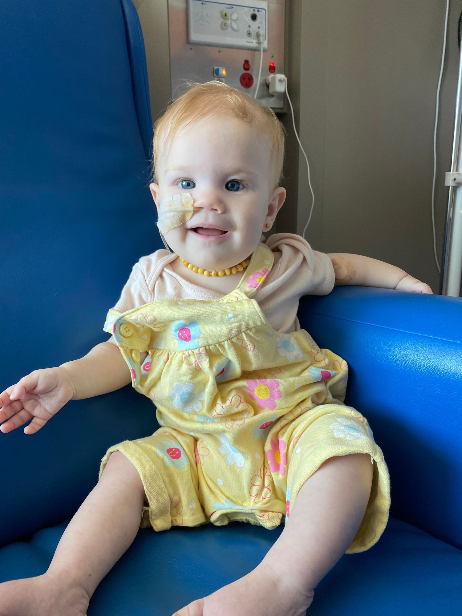 Smiling baby girl in yellow floral romper with a nasal tube sitting in a blue hospital chair.