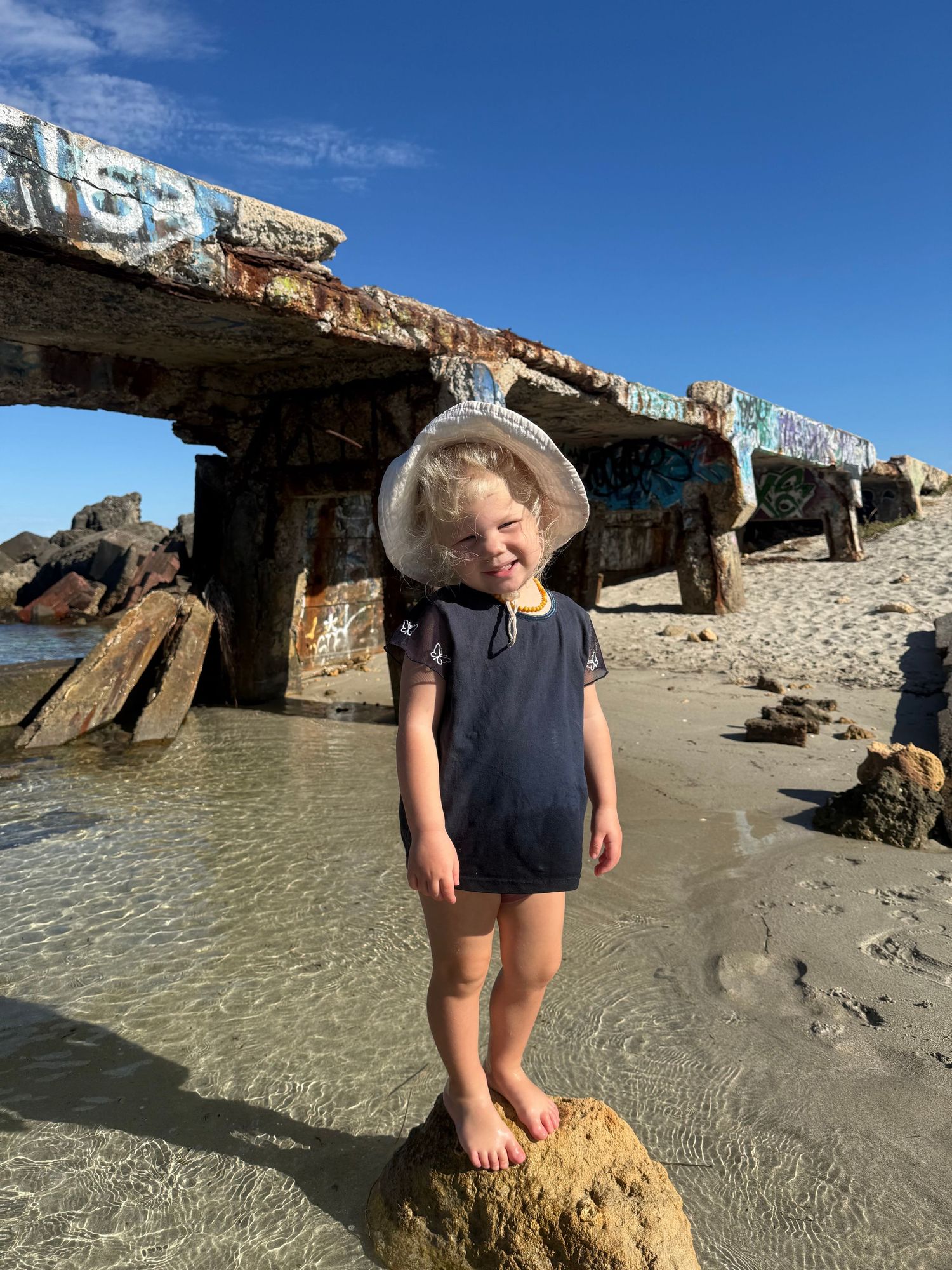 Happy toddler standing on a rock at a graffiti-covered beach ruin wearing a sun hat and navy swimsuit.