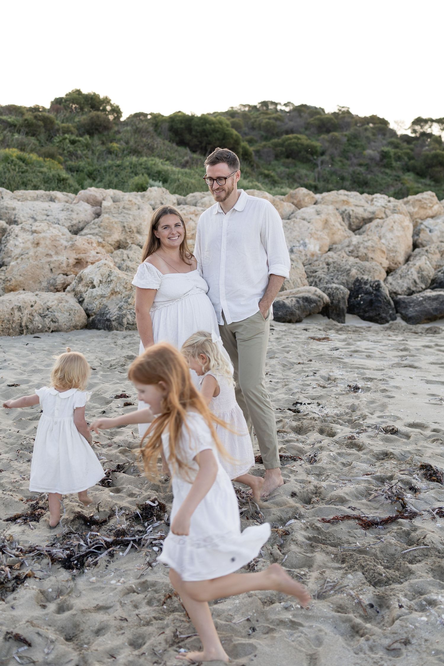 Pregnant woman and man smile as two young girls in white dresses play on a rocky beach at sunset. Taken by Golden Hour Perth at Cape Peron