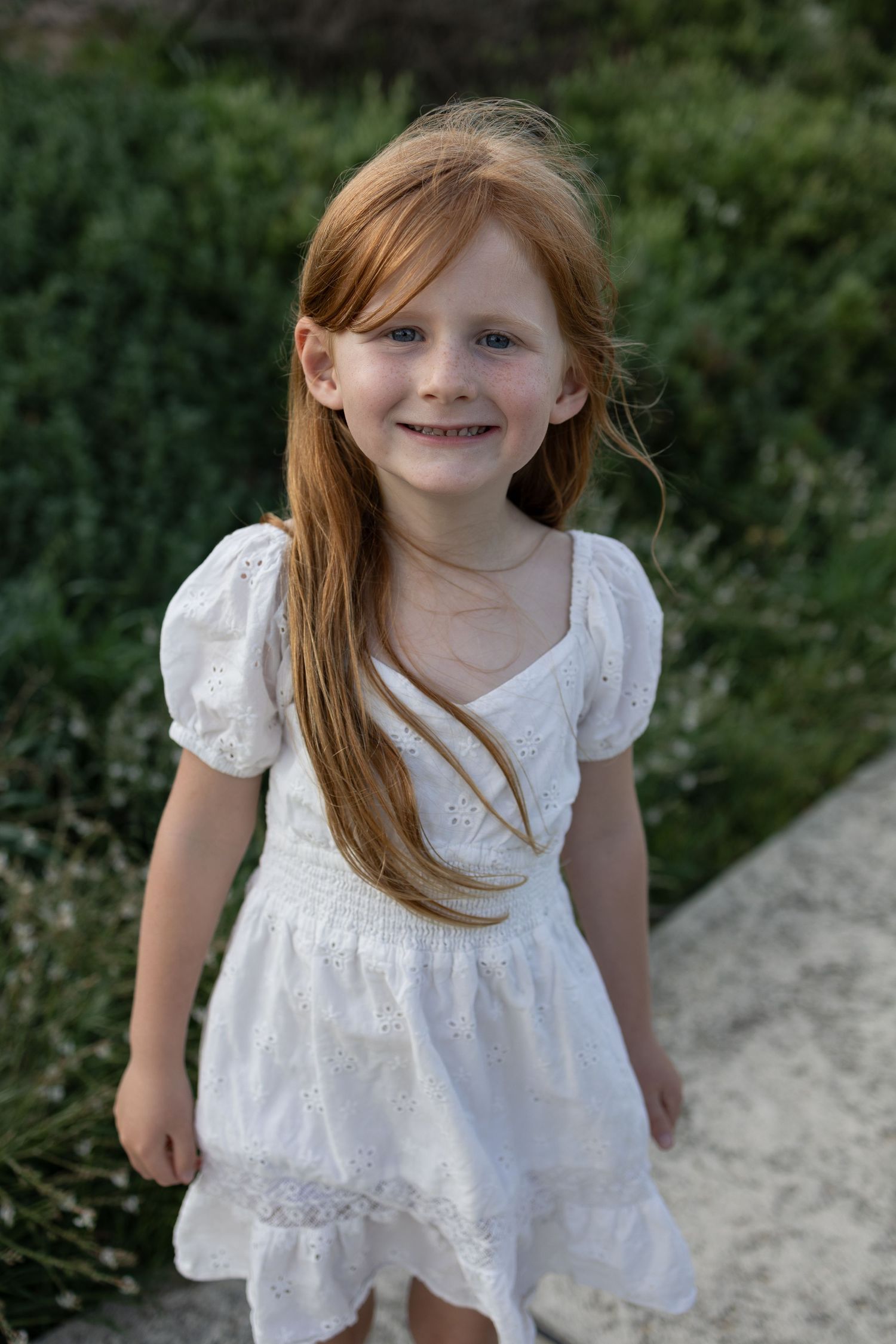 Young girl with long red hair smiling in a white dress, standing outdoors near green shrubs. Taken by Golden Hour Perth  at Cape Peron